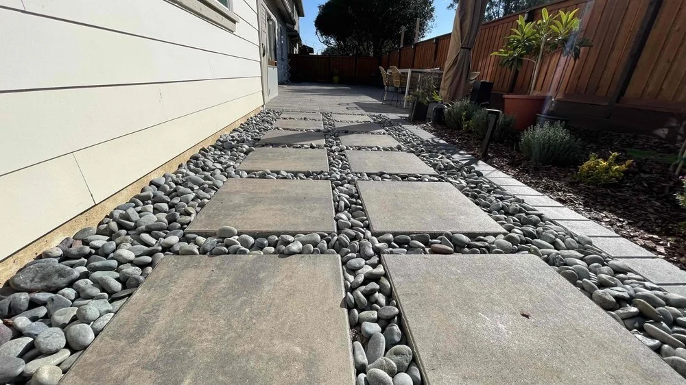 Residential backyard pathway with large concrete stepping stones and smooth pebbles in between, bordered by a white house wall on the left and a wooden fence with plants on the right, under a blue sky with trees in the background.