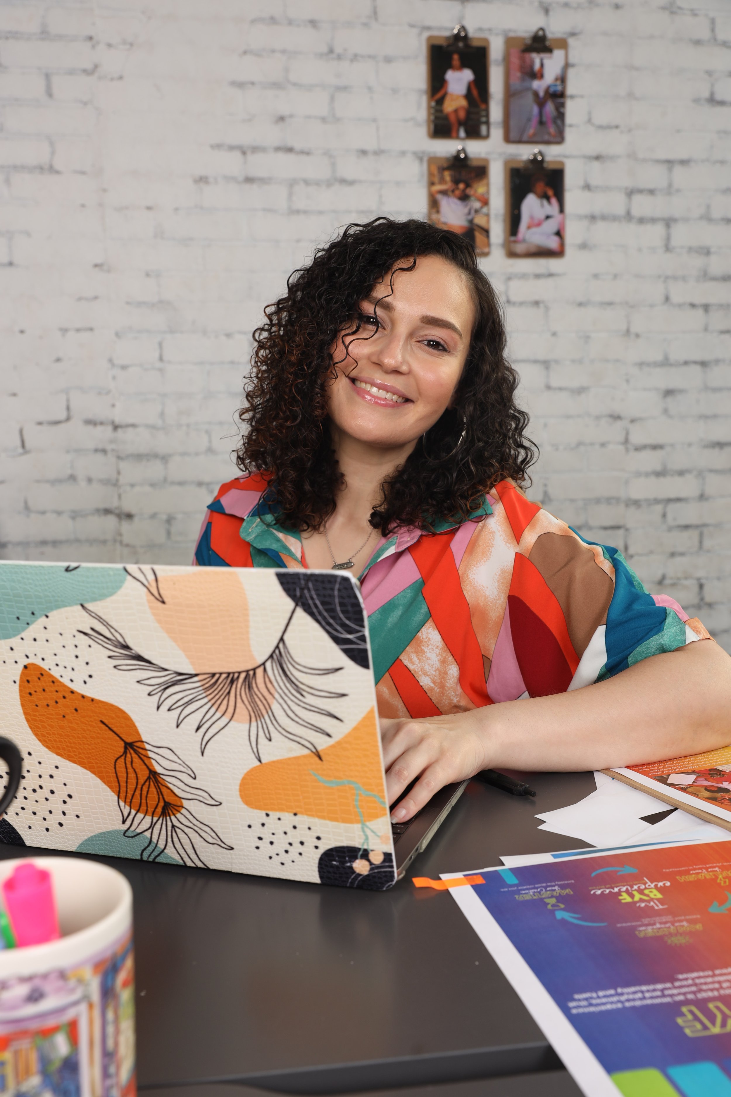 A woman with curly hair, smiling, working on a laptop at a desk with colorful artwork on her laptop and various papers and objects around her, gold clips with photographs hanging on a white brick wall in the background.