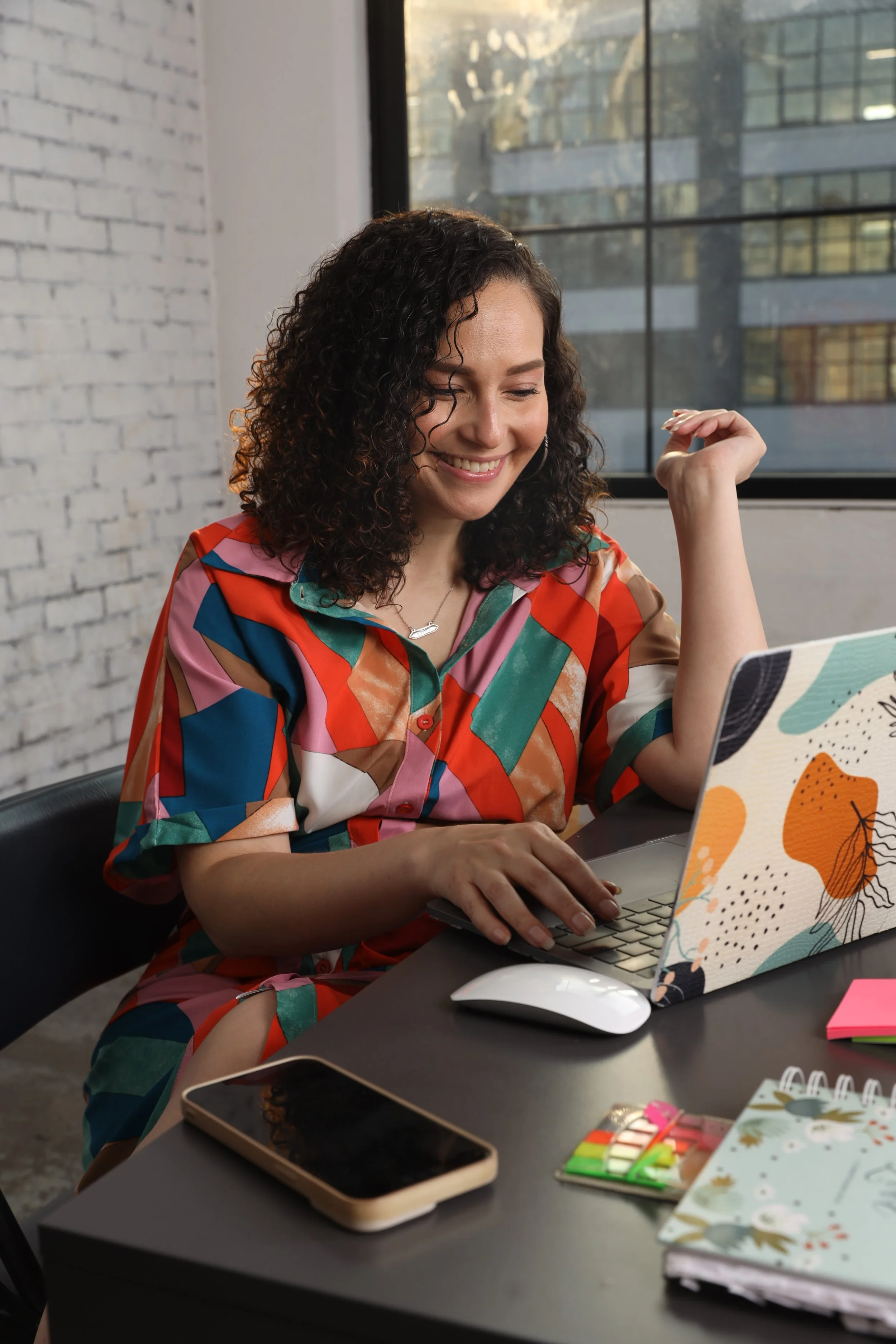 A woman with curly hair wearing a colorful, patterned shirt, smiling while working on a laptop at a desk in a modern office with large windows and a white brick wall.