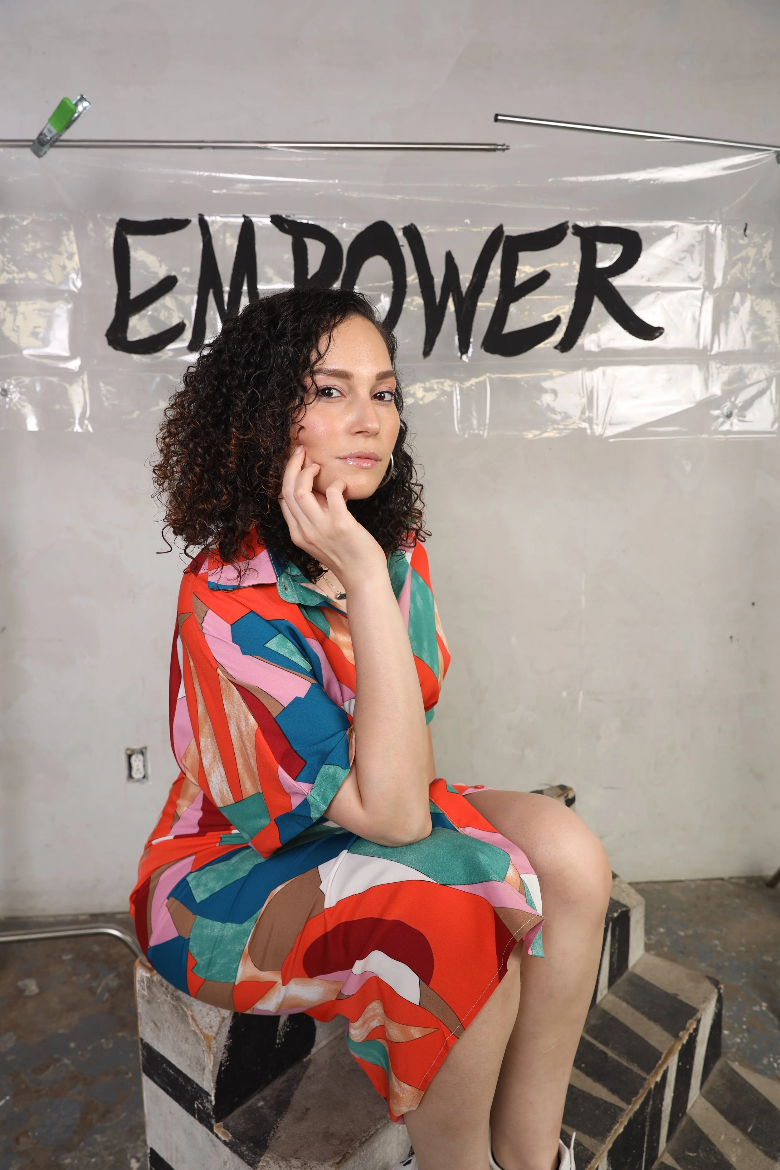 Woman seated on a painted block, wearing a multicolored dress, with a backdrop of a clear plastic banner that has the word 'EMPOWER' written in bold black letters.