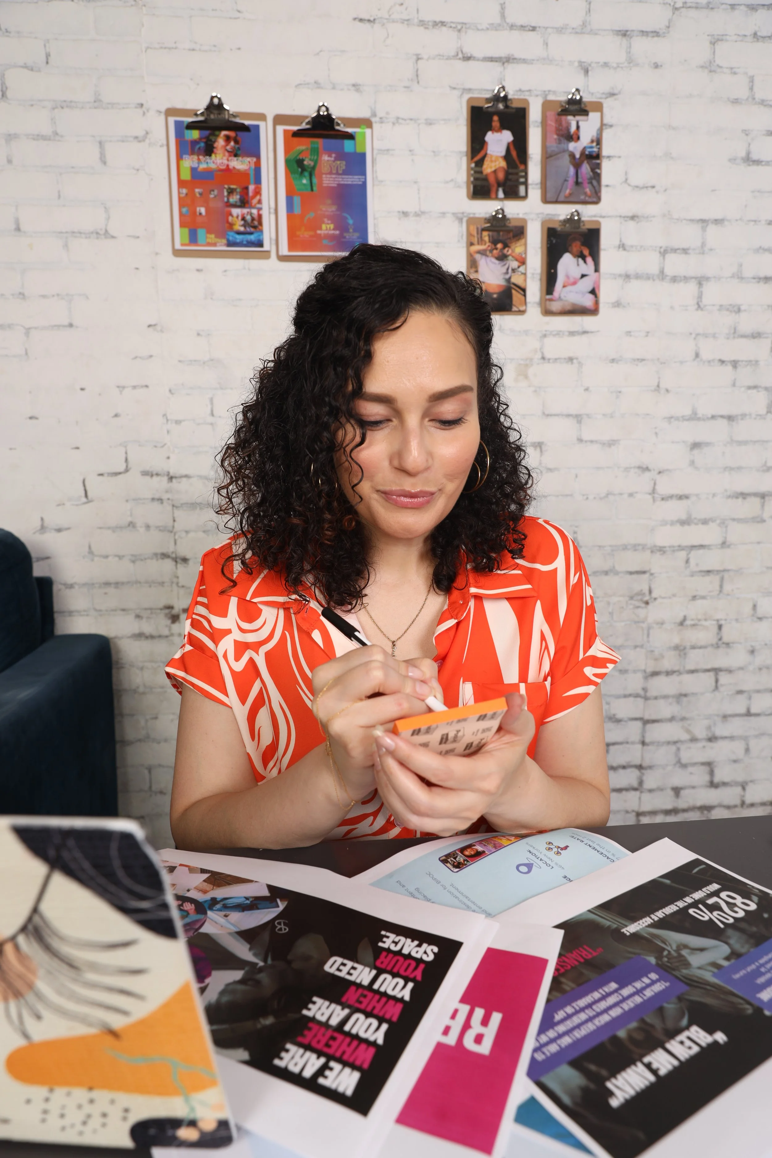 A woman with curly dark hair, wearing a red patterned blouse, is sitting at a table, writing or coloring in a small notebook or journal. There are various flyers, papers, and magazines spread out on the table in front of her. The background features a white brick wall with several photographs and posters clipped to it.