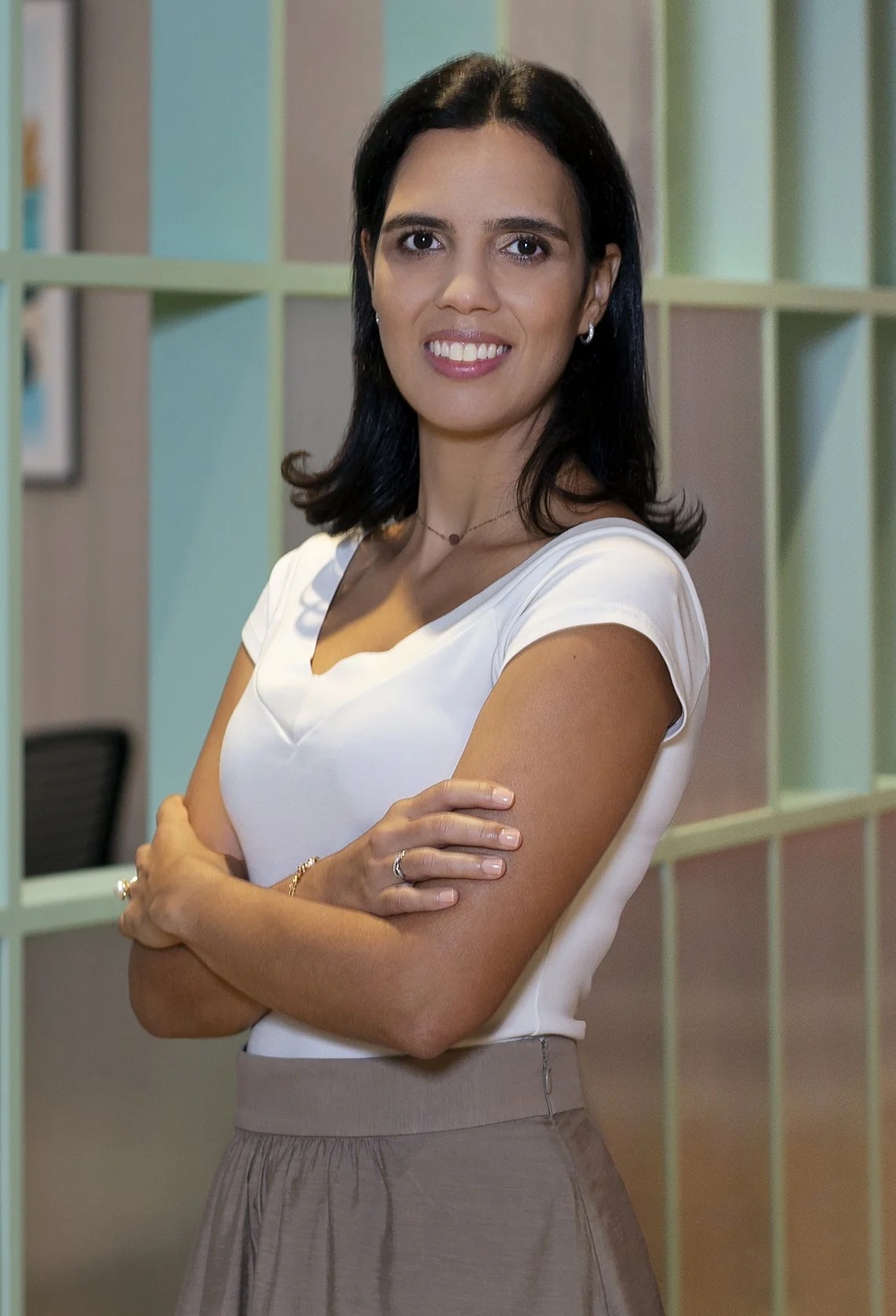 Mulher de cabelo preto e curto, vestindo camiseta branca, está sorrindo e com os braços cruzados. Fundo com divisórias verdes.