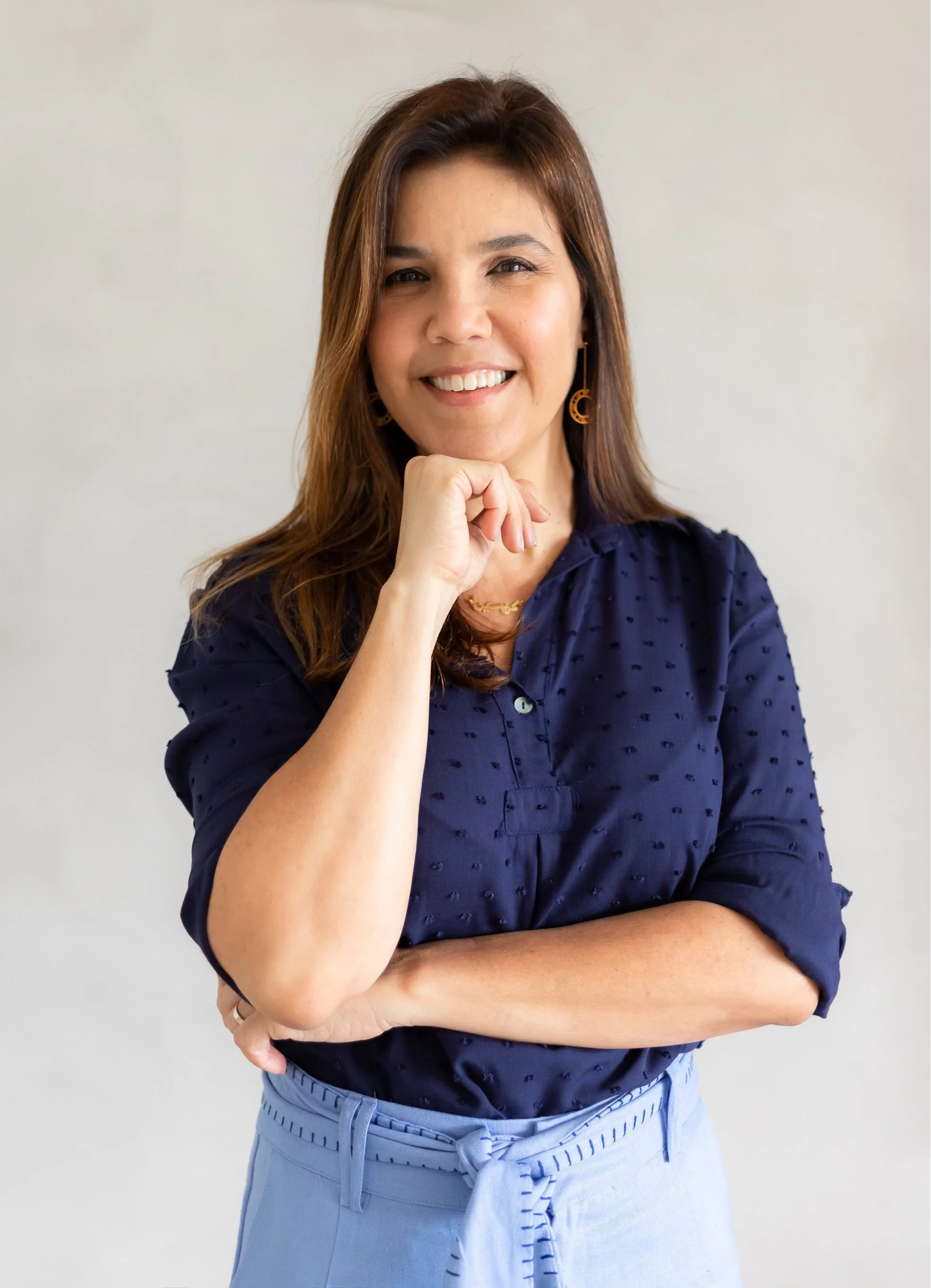 Mulher sorridente com cabelo castanho, camisa azul marinho, segurando o queixo com a mão, fundo cinza claro.