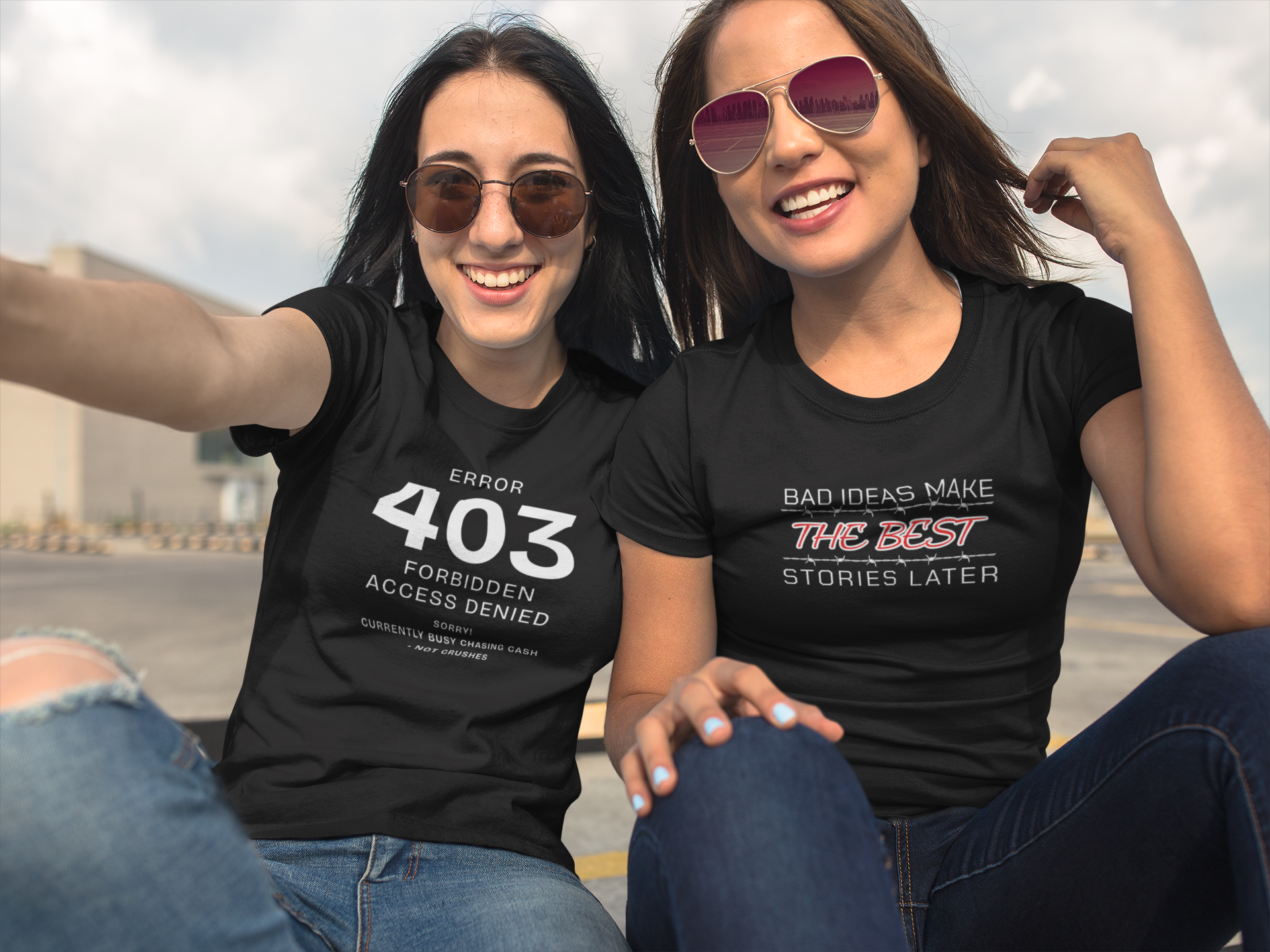 Two women taking a selfie outdoors, both wearing black T-shirts with humorous text, and sunglasses. They are smiling and sitting on a flat rooftop or parking lot with a cloudy sky in the background.