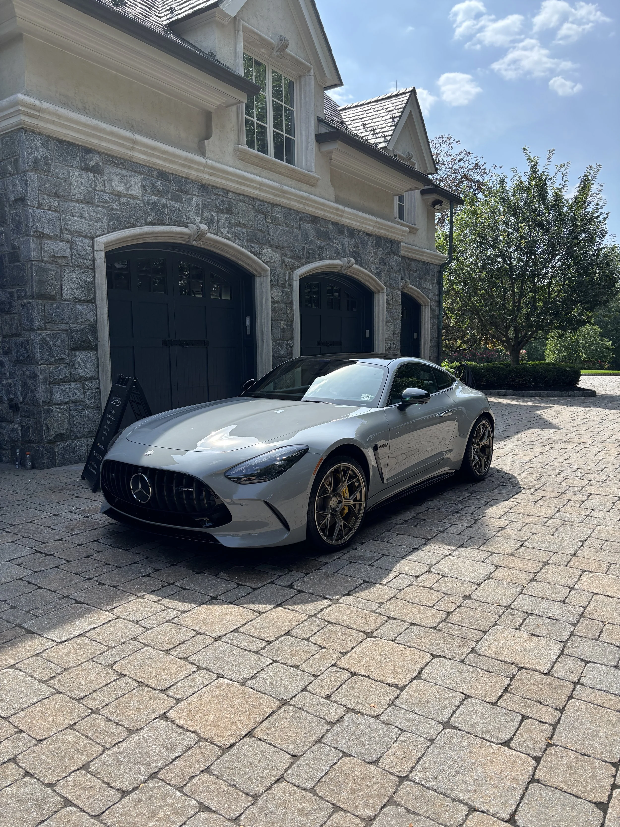 A silver Mercedes-Benz AMG sports car parked on a stone driveway in front of a large house with stone and beige exterior.