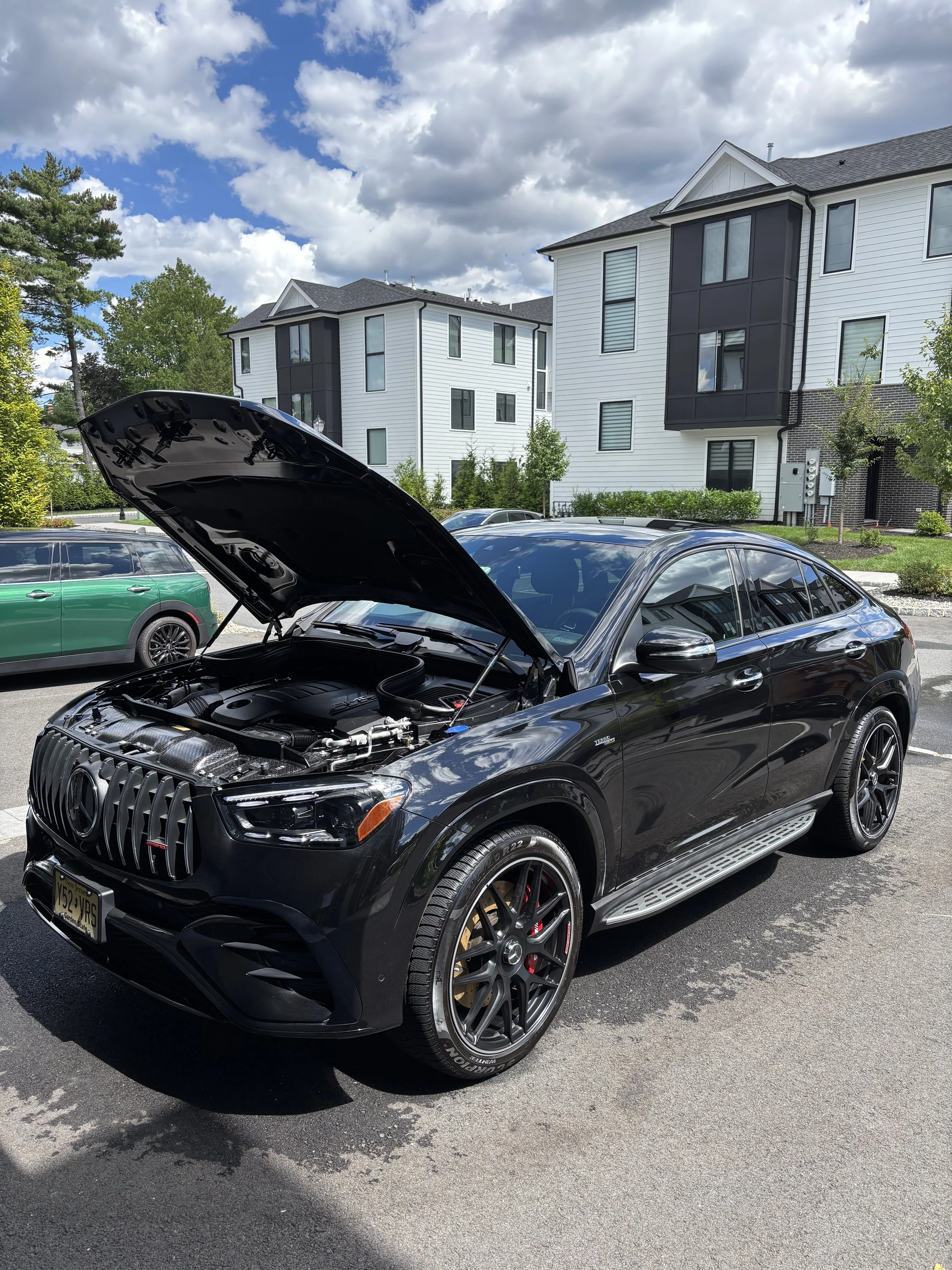 A black Mercedes-Benz SUV parked in a parking lot with its hood open, revealing the engine. In the background, there are residential buildings, greenery, and another green vehicle.