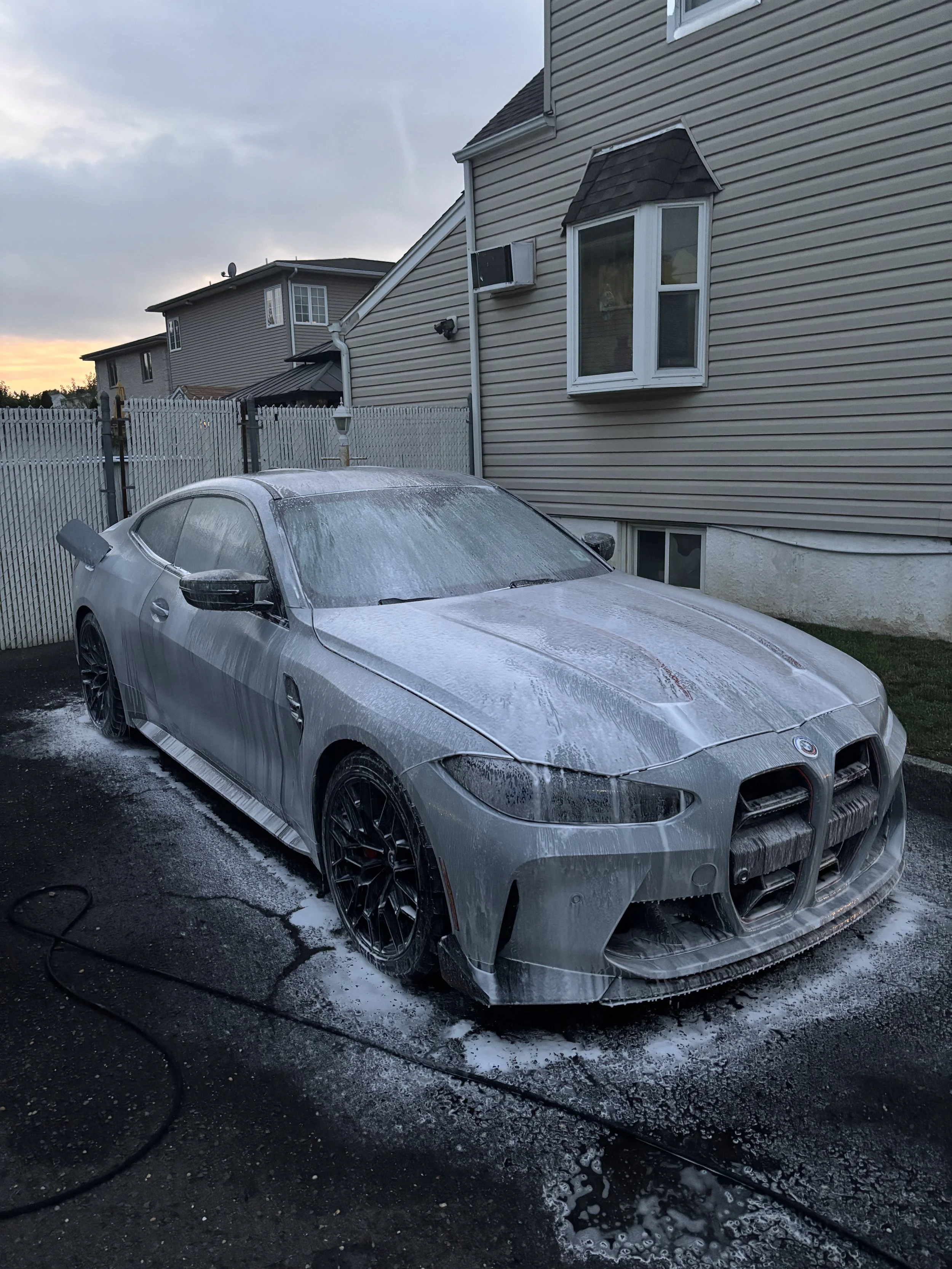 A silver sports car is being washed and covered with soap suds in a driveway, with a residential house and neighboring houses in the background.