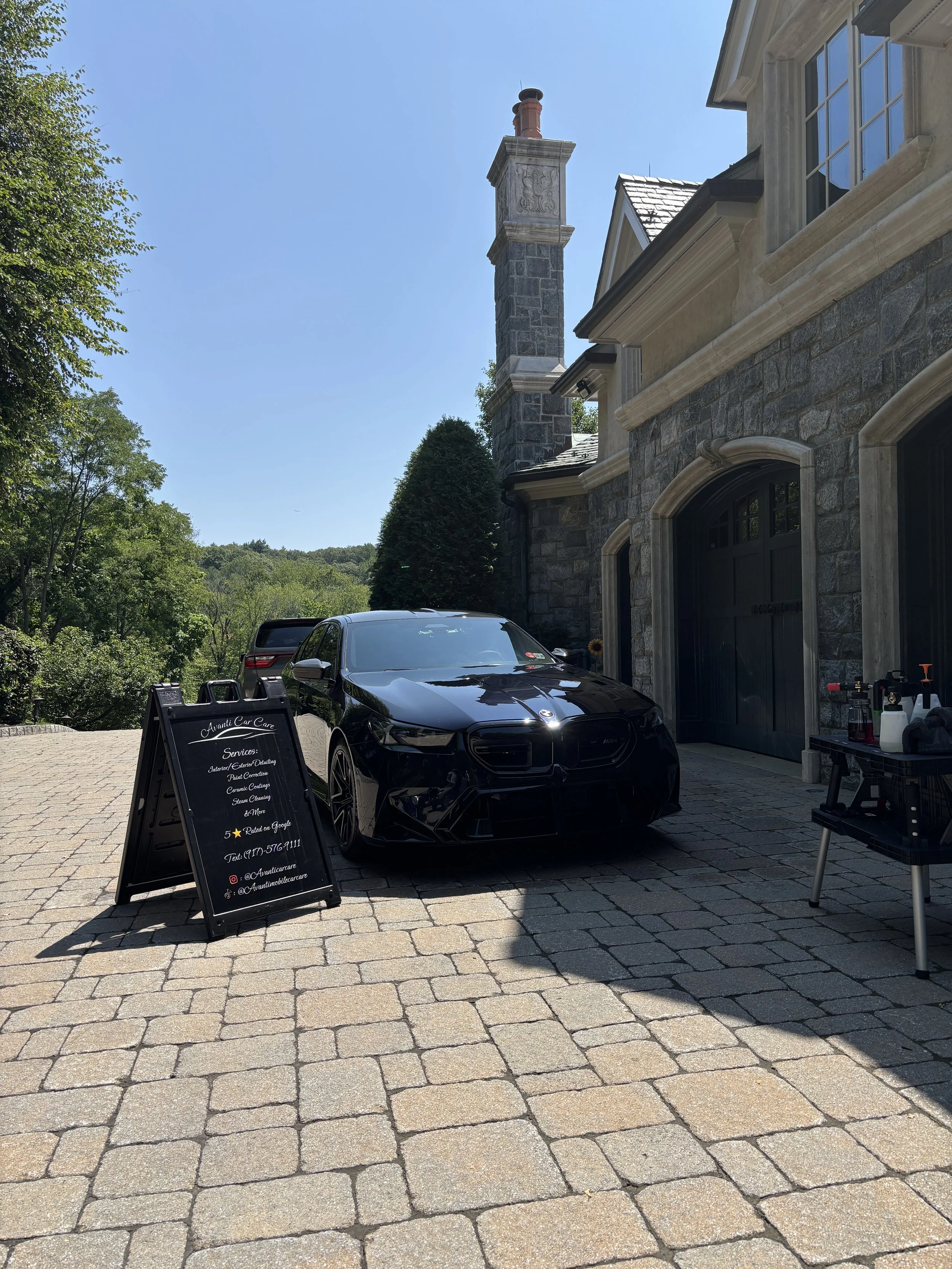 Black car parked on driveway in front of a stone house with a signboard for auto detailing services.