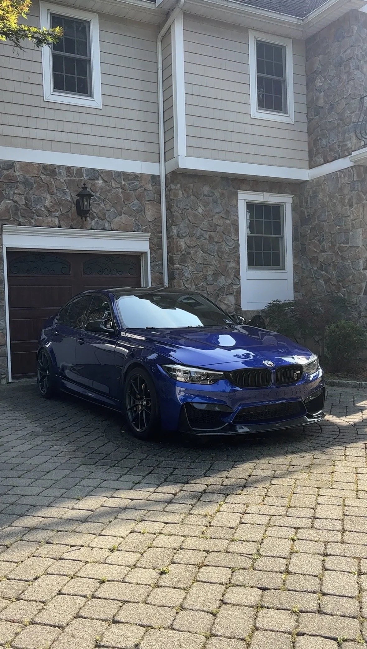 A blue BMW sedan parked on a cobblestone driveway in front of a house with stone and beige siding, white window frames, and a garage door.