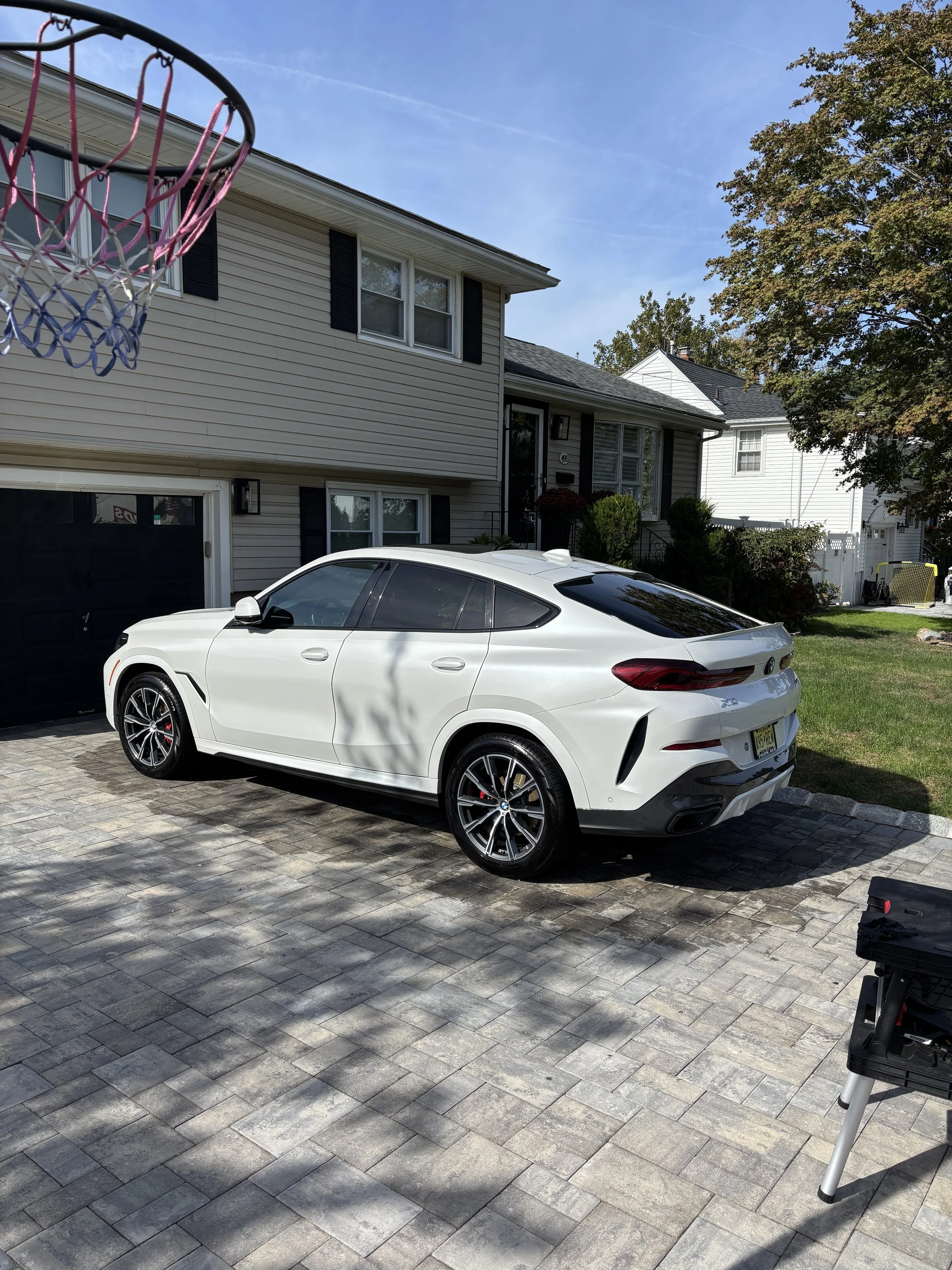 A white modern SUV parked on a stone-paved driveway in front of a beige house with black shutters under a blue sky. A basketball hoop is partially visible in the top left corner.
