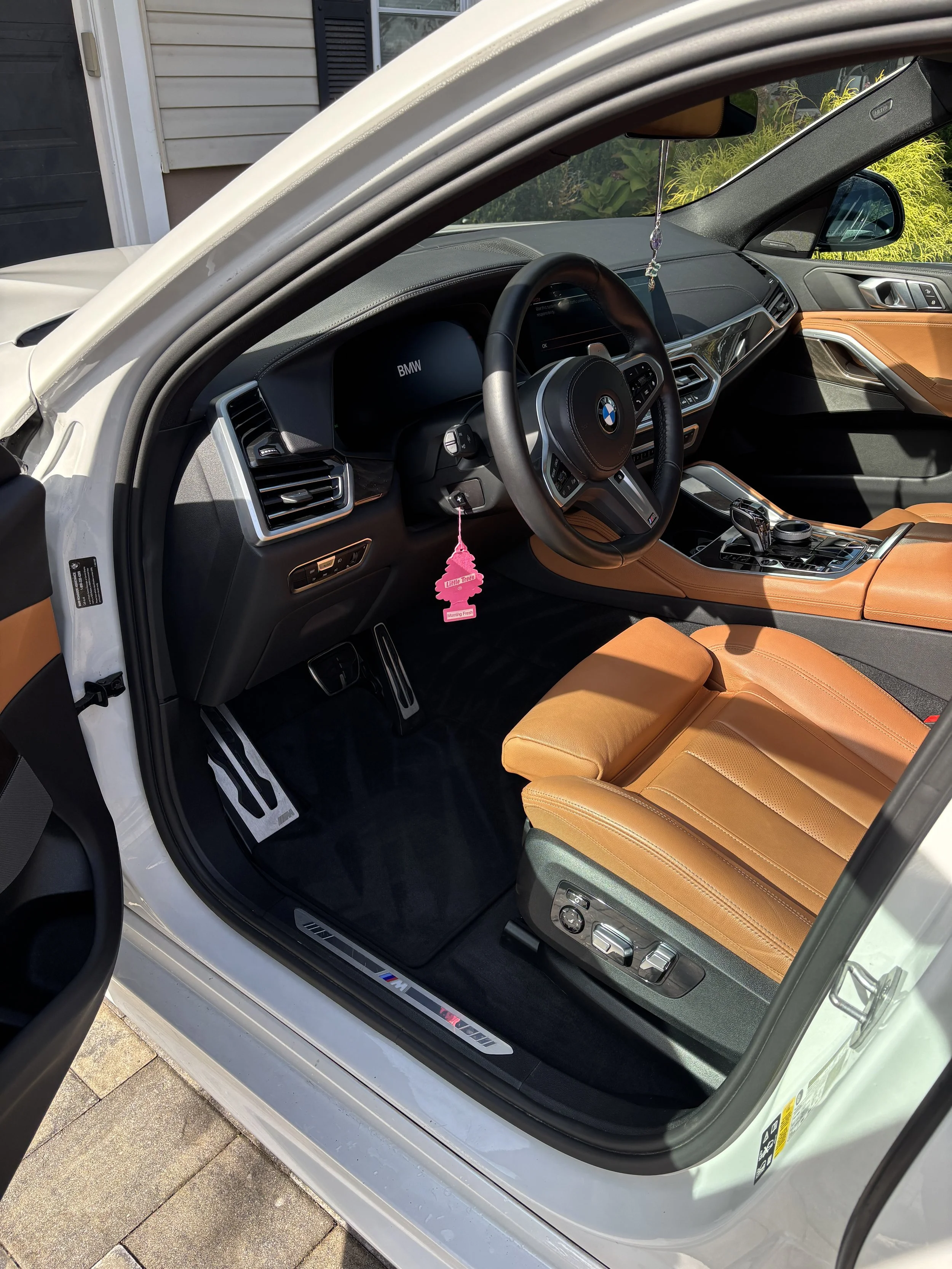 Interior of a white BMW car, showing tan leather seats, black dashboard with digital displays, and a steering wheel with controls. The side door has a wood trim, and a pink tree-shaped air freshener hangs from the rearview mirror.