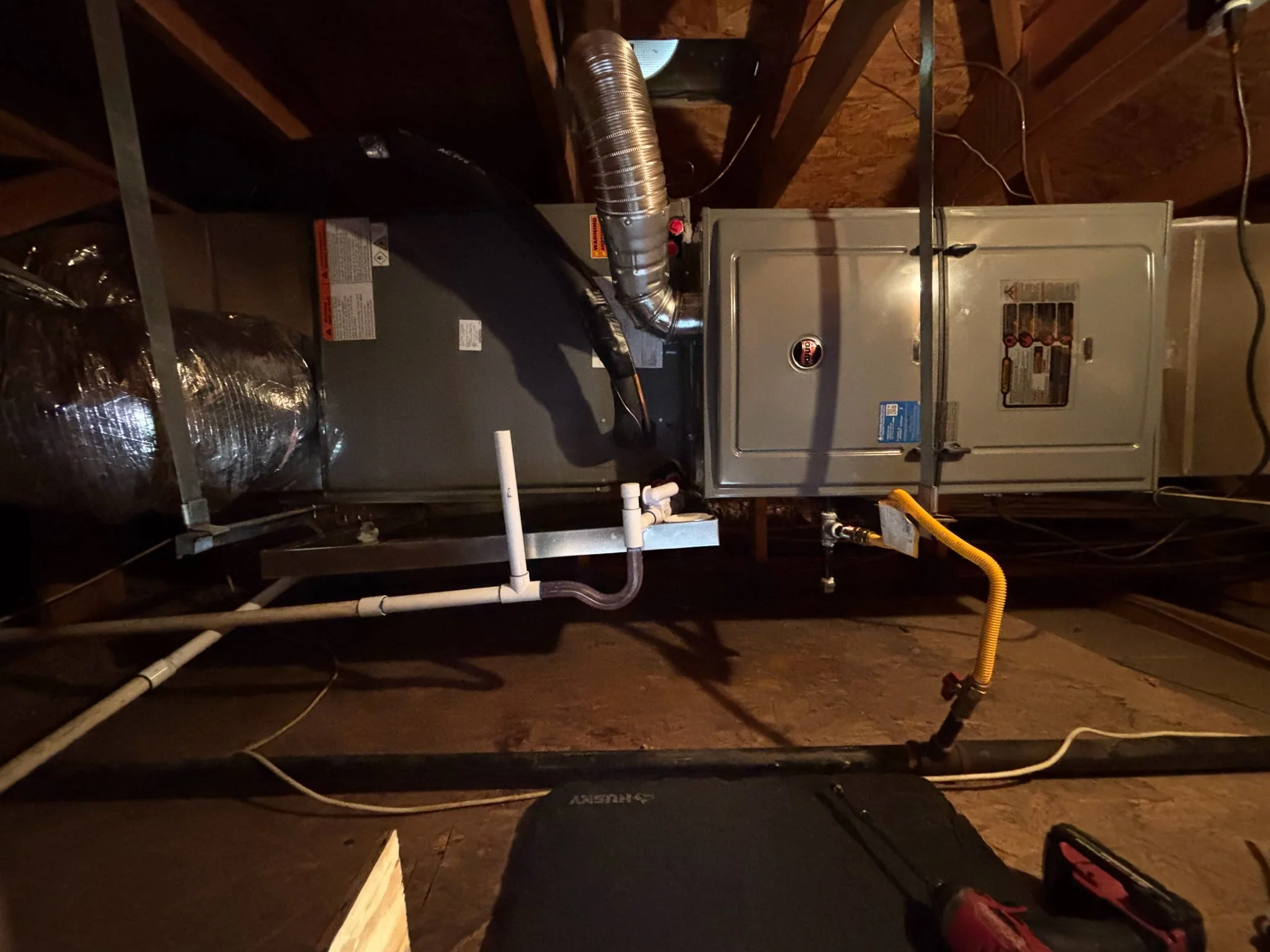 An HVAC furnace system installed in a wooden ceiling space with various ducts, pipes, and wiring.