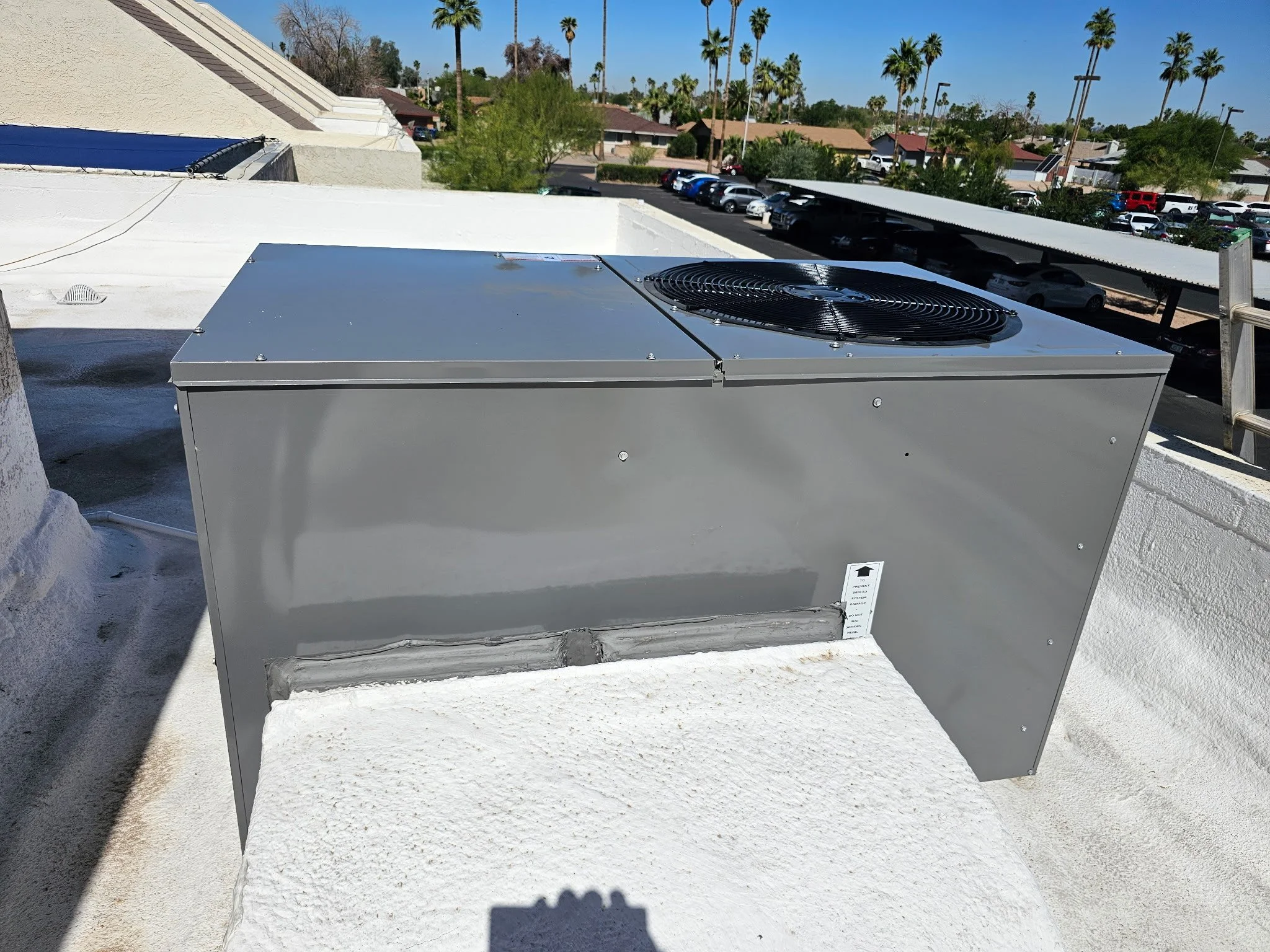 A rooftop air conditioning unit with a large fan on top, mounted on a white textured surface with a parking lot and palm trees in the background.