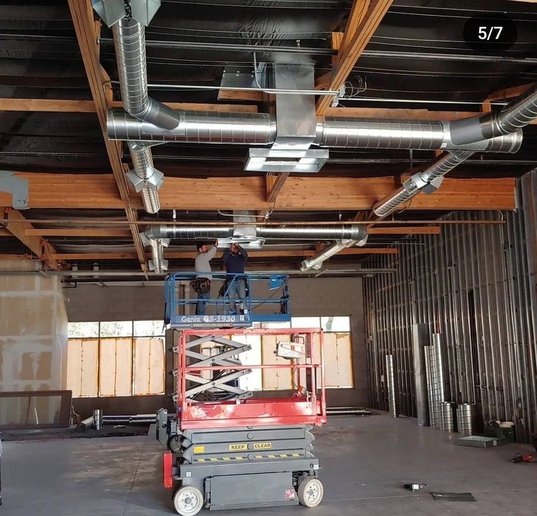 Two workers on a blue scissor lift installing or inspecting ceiling ductwork in an unfinished building with exposed wooden beams and metal wall studs.