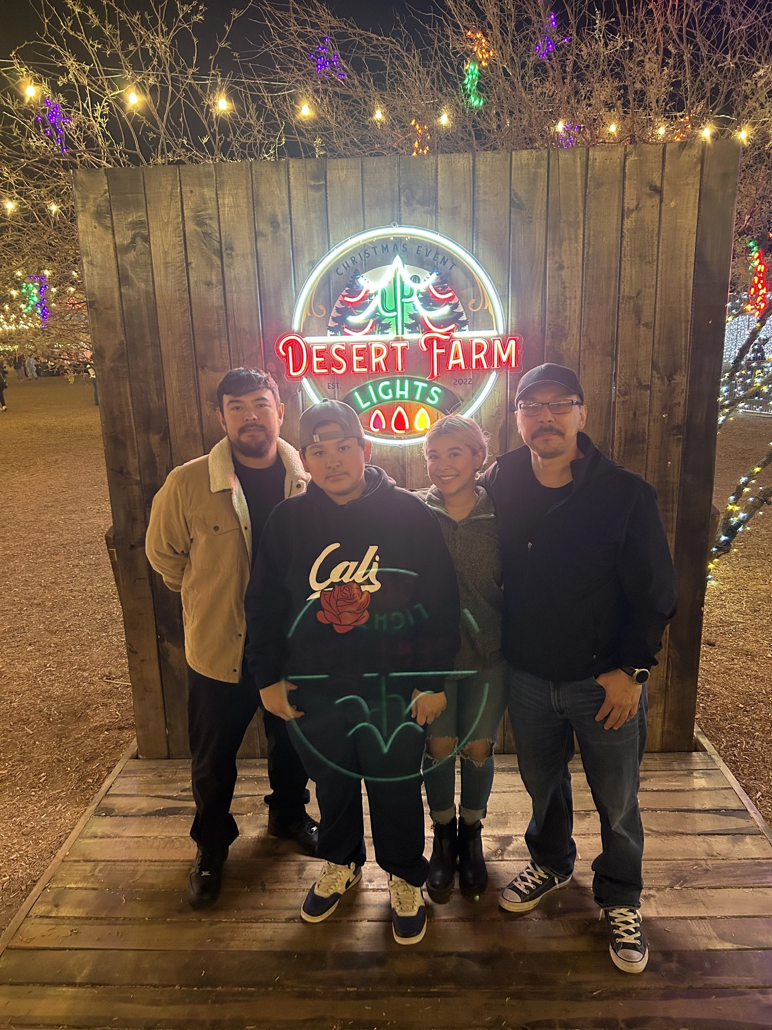 Four people standing in front of a wooden backdrop with a neon sign that reads "Desert Farm Lights," at a Christmas event at night. The sign features a Christmas tree and holiday colors. Two men and two women, all smiling.