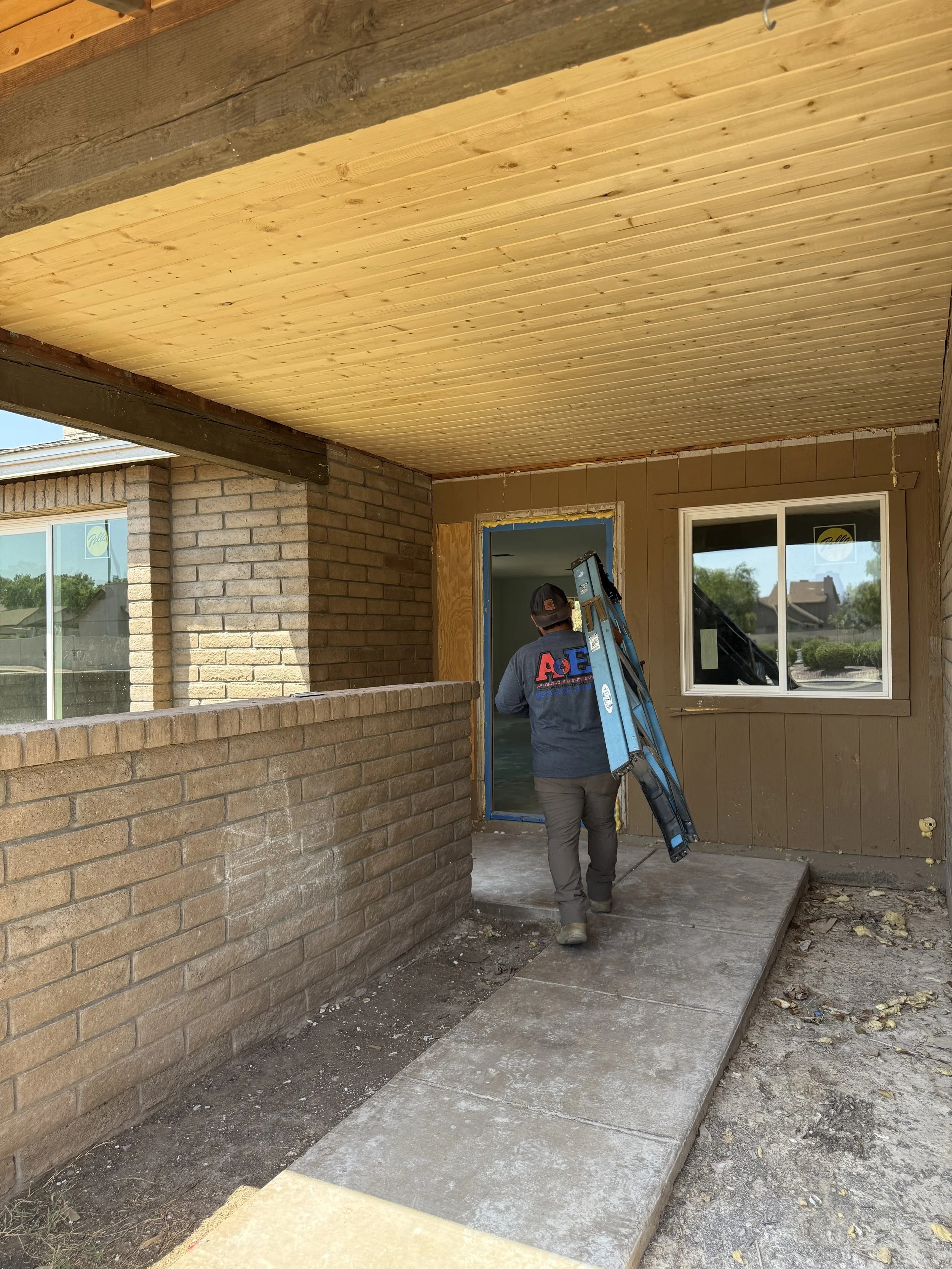 A construction worker walking on a concrete porch carrying a ladder into a house under renovation. The porch has a brick wall on one side, a wooden ceiling, and a door frame being prepped for installation. The worker is wearing a dark sweatshirt with