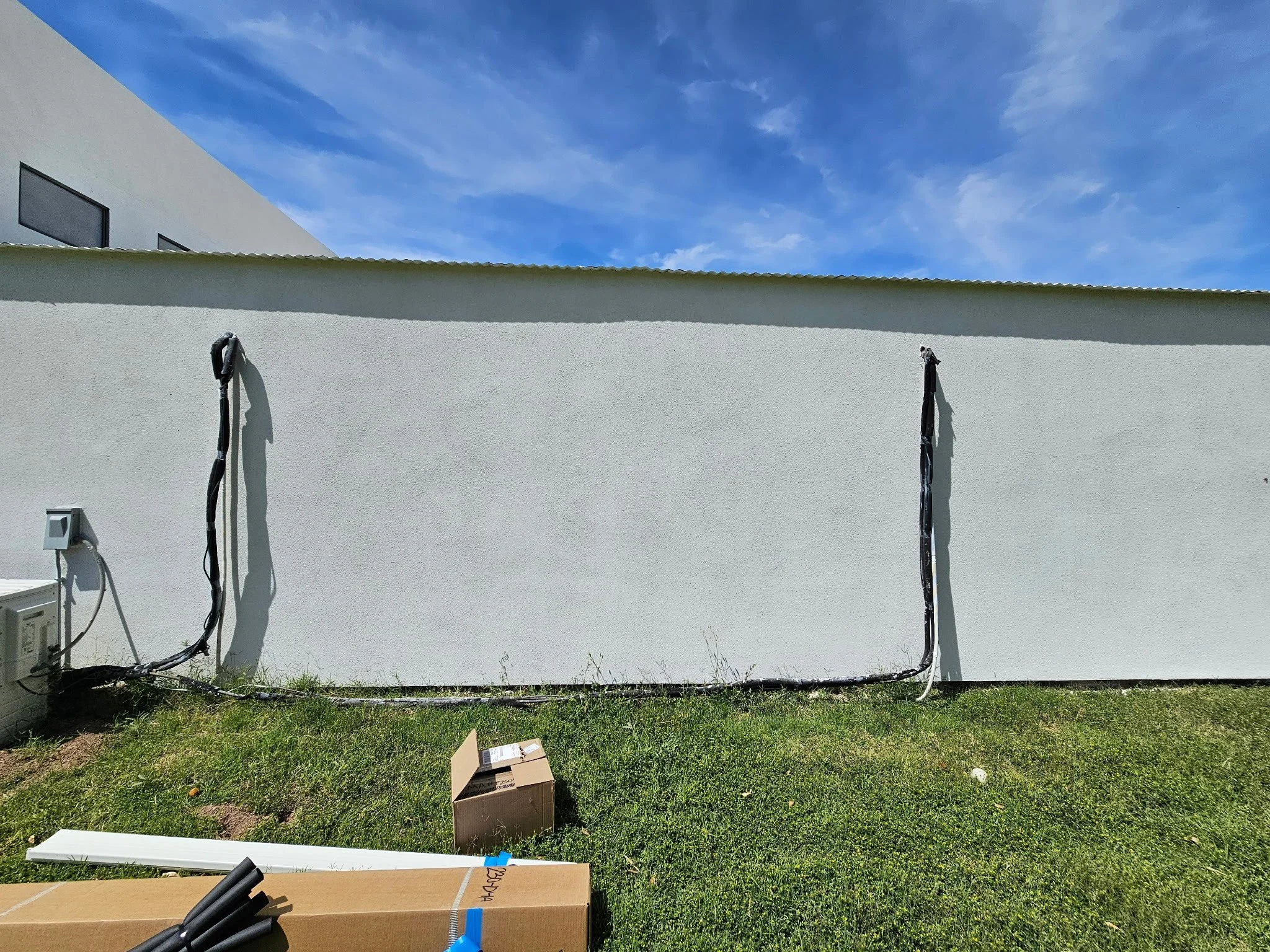 Blank white exterior wall of a building with black electrical cables mounted on it, on a grassy ground under a blue sky with some clouds, with a cardboard box and some black pipes on the ground.