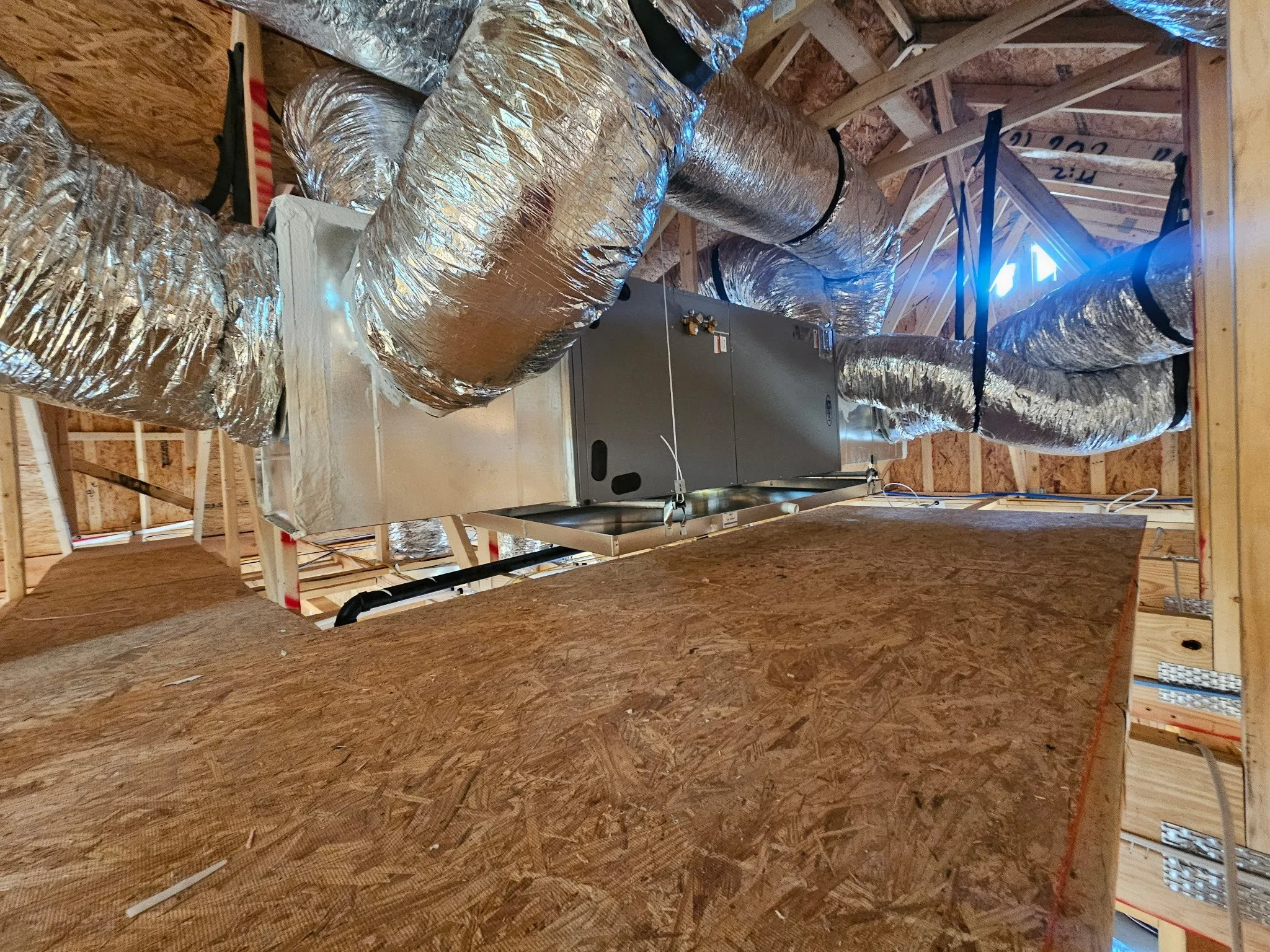 View of a building's attic space showing metallic HVAC ductwork, a large metal air handler, wooden framing, and insulation panels.