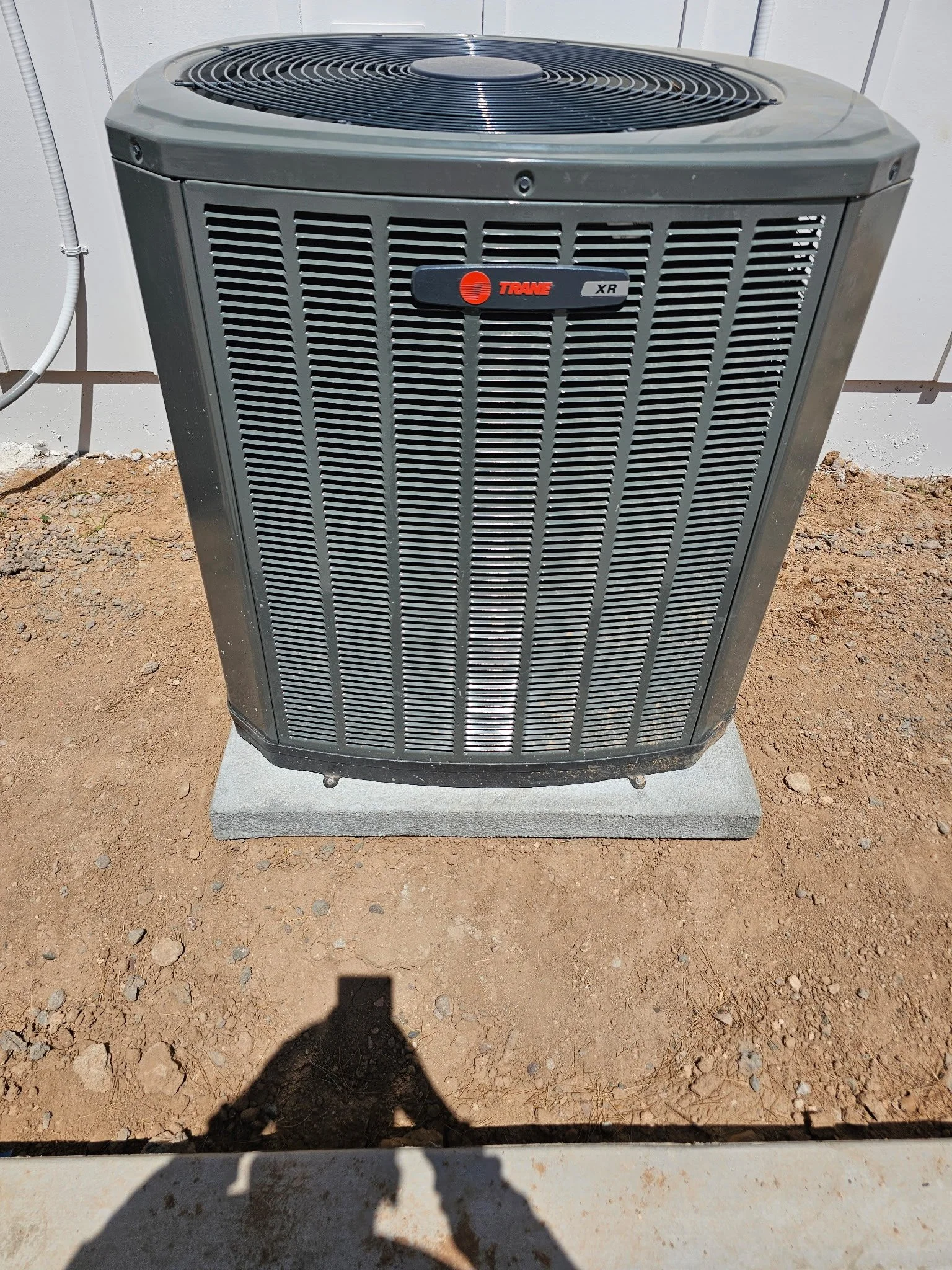 An outdoor air conditioning unit with a gray metal grille, sitting on a concrete slab on dirt ground, with a shadow of a person taking the photo visible on the ground.