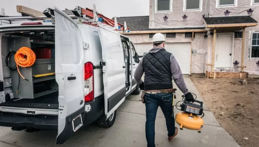 A construction worker walks away from a white service van carrying a portable air compressor, on a residential construction site with a partially built house in the background.