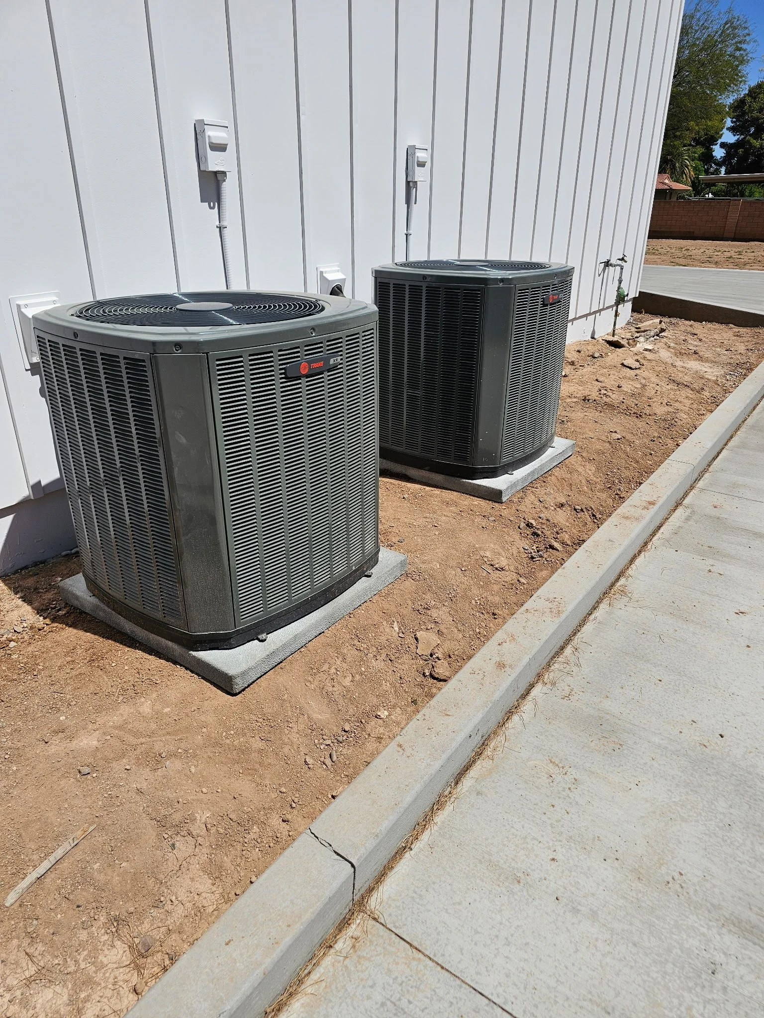 Two outdoor air conditioning units installed side by side on concrete pads next to a light-colored building with white siding. The units are situated on a dirt area, with a sidewalk running along the edge.