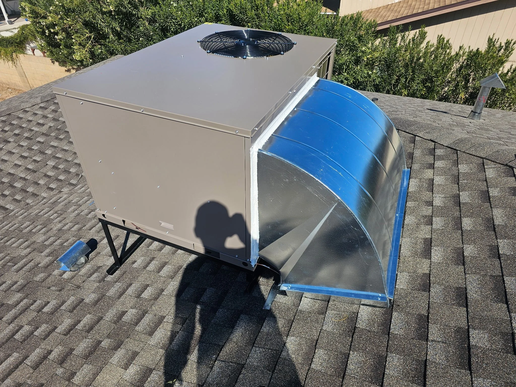 A rooftop HVAC unit with a fan on top and a curved duct connected to the side, installed on asphalt shingles.