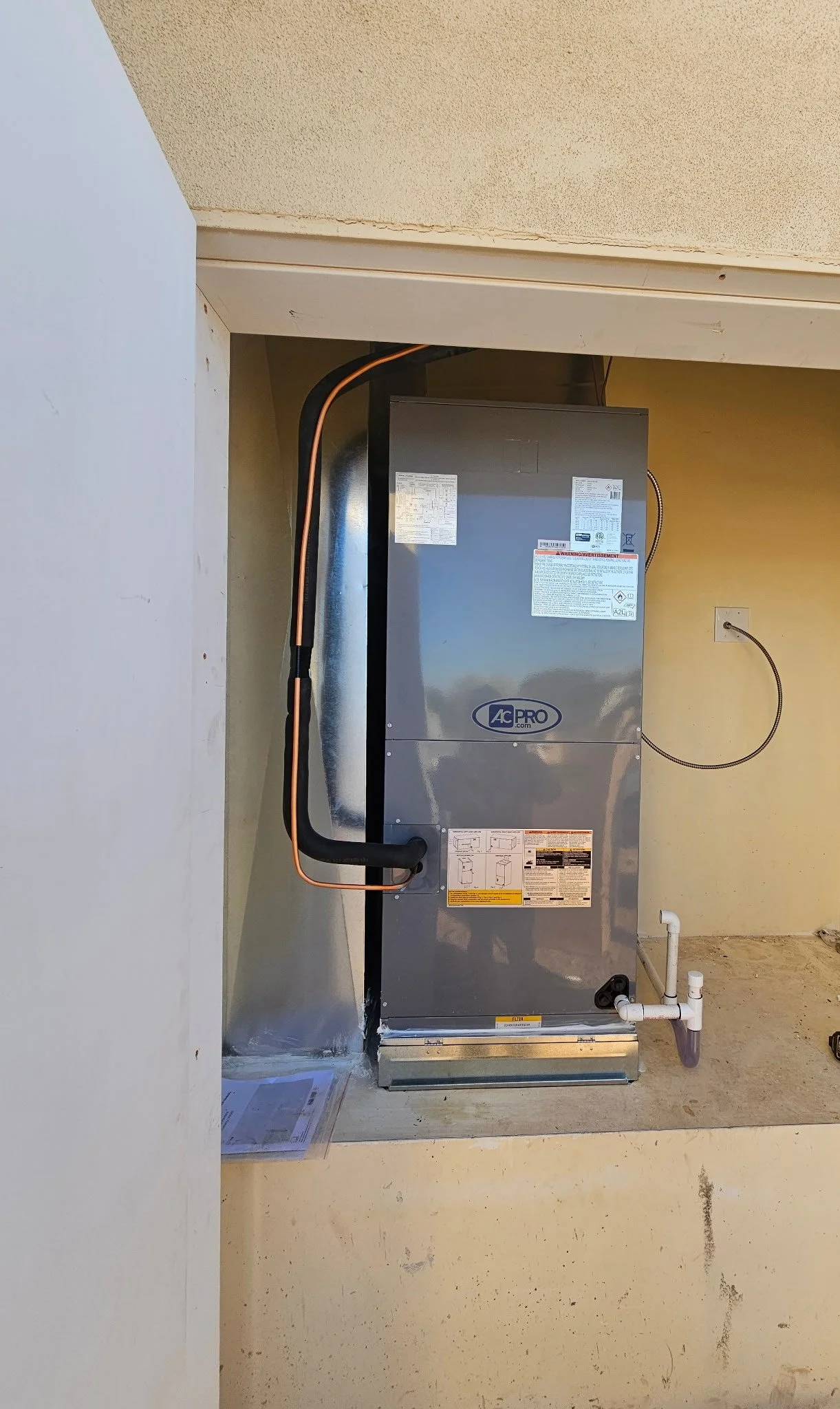 Installation of an AC Pro air conditioning unit on a concrete slab with piping and electrical connections, next to a wall with outlet.