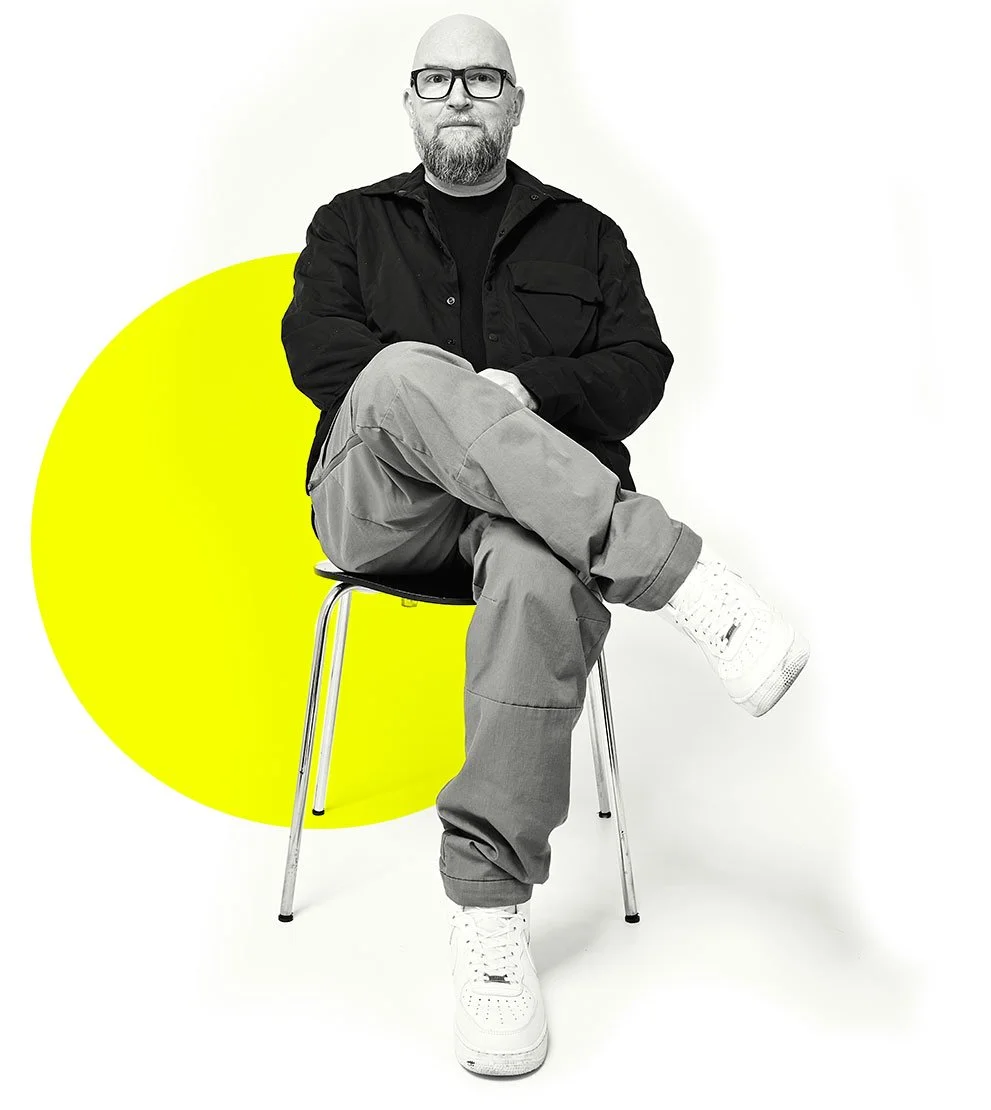 Mann mit Brille, grauem Bart, schwarzer Jacke, grauen Hosen und weißen Turnschuhen sitzt auf einem Stuhl vor weißem Hintergrund mit gelbem Kreis im Hintergrund.