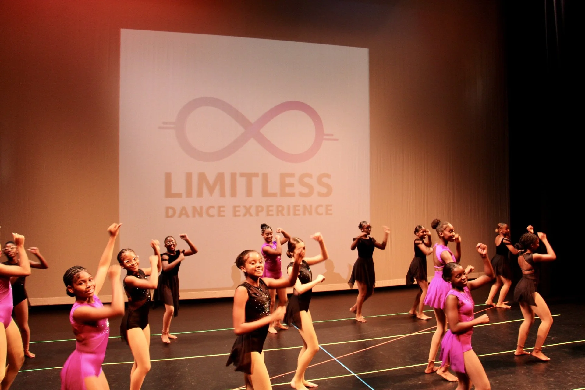 Group of young girls dancing on stage during a performance with a large screen in the background displaying the logo and name 'Limitless Dance Experience'.