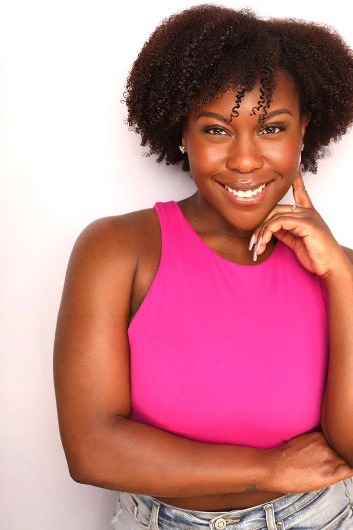 A young woman with dark curly hair, smiling, wearing a bright pink sleeveless top, earrings, and light-colored jeans, standing against a plain white background.