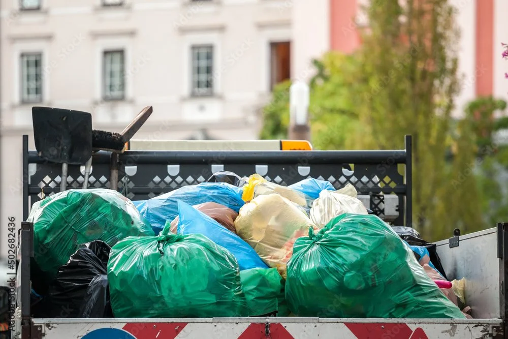 A truck filled with green, black, and beige garbage bags parked outdoors with buildings and trees in the background.