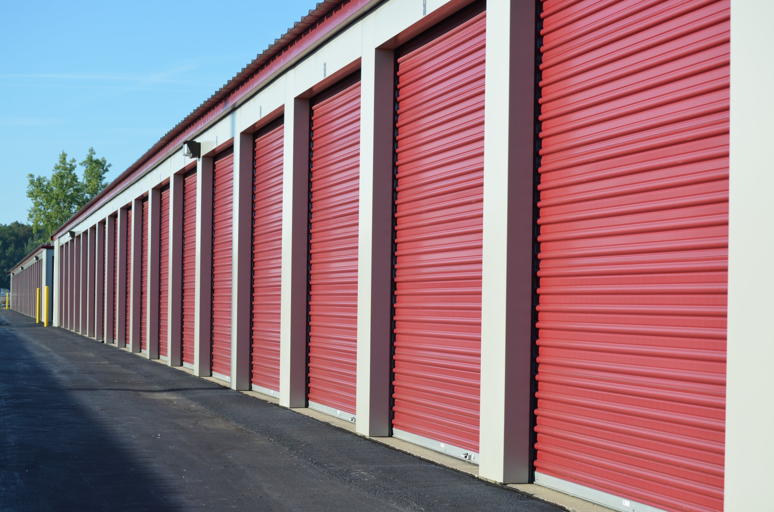 A row of storage unit doors with red metal roll-up doors and white vertical supports, under a partly cloudy sky.