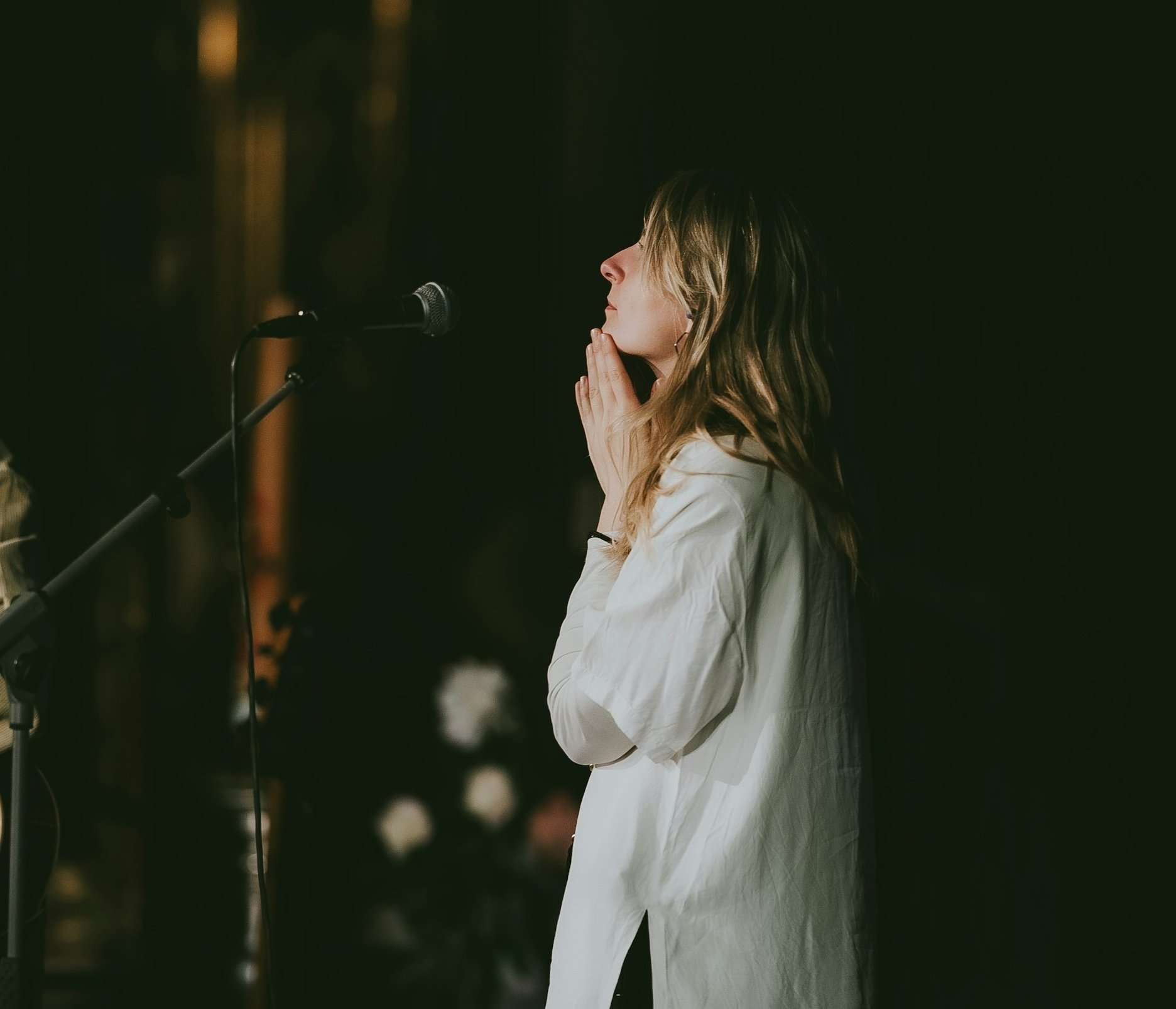 A woman with long wavy hair and a white shirt standing in front of a microphone with her hands pressed together near her face, eyes closed, in a dark setting.