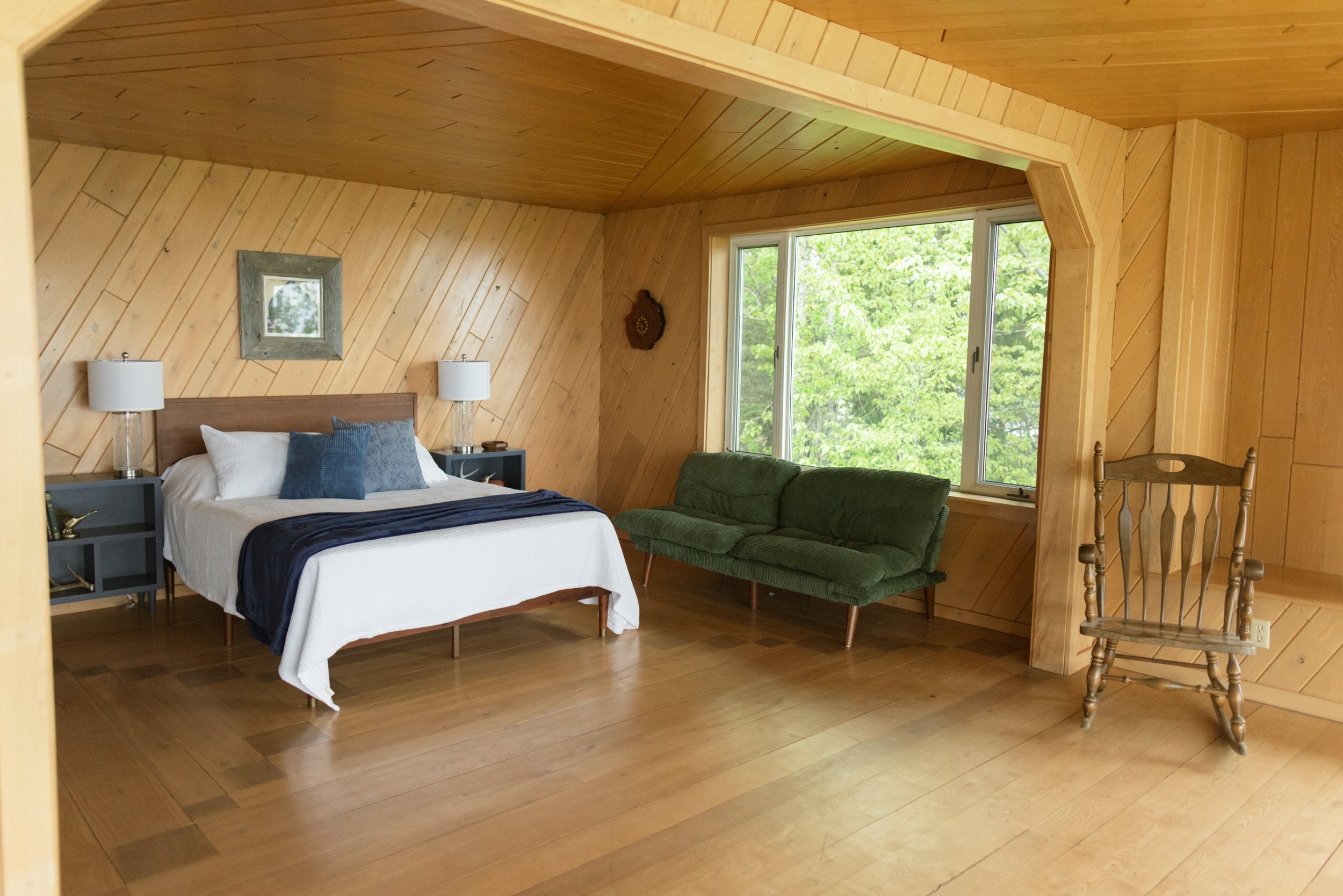View of a cozy bedroom with wooden wall paneling and ceiling, featuring a bed with white and blue bedding, two bedside lamps, a window with greenery outside, a green vintage-style couch, and a wooden chair.