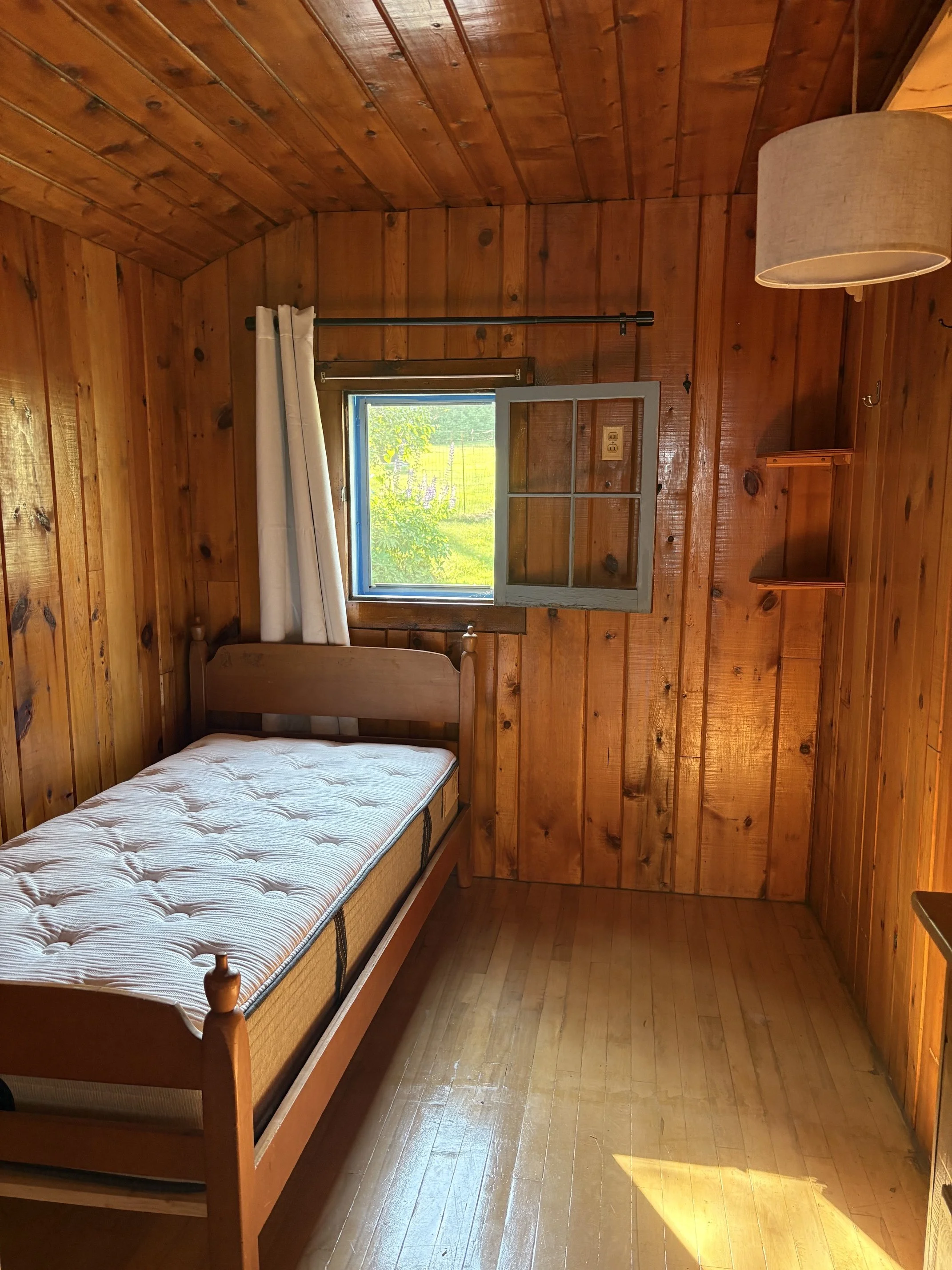 A small bedroom with wooden walls and ceiling, a single bed with a mattress, a small window with white curtains, and a beige lampshade hanging from the ceiling.