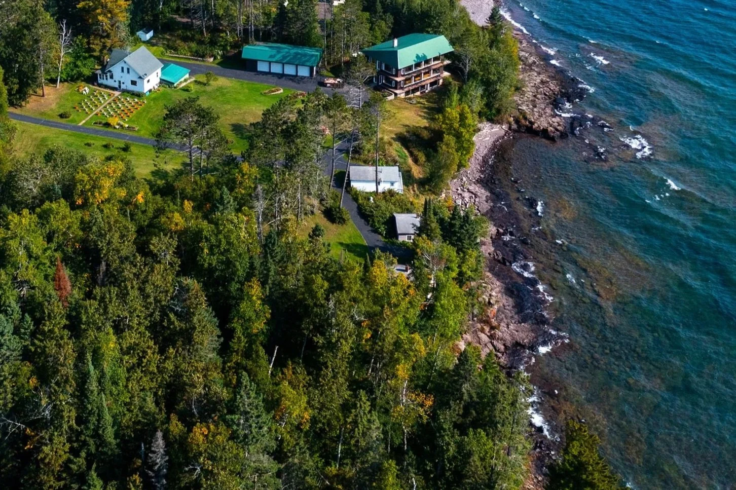 Aerial view of a coastal landscape with a forested area, a road running parallel to the shoreline, and several houses with green roofs, alongside the rocky shoreline and blue water.