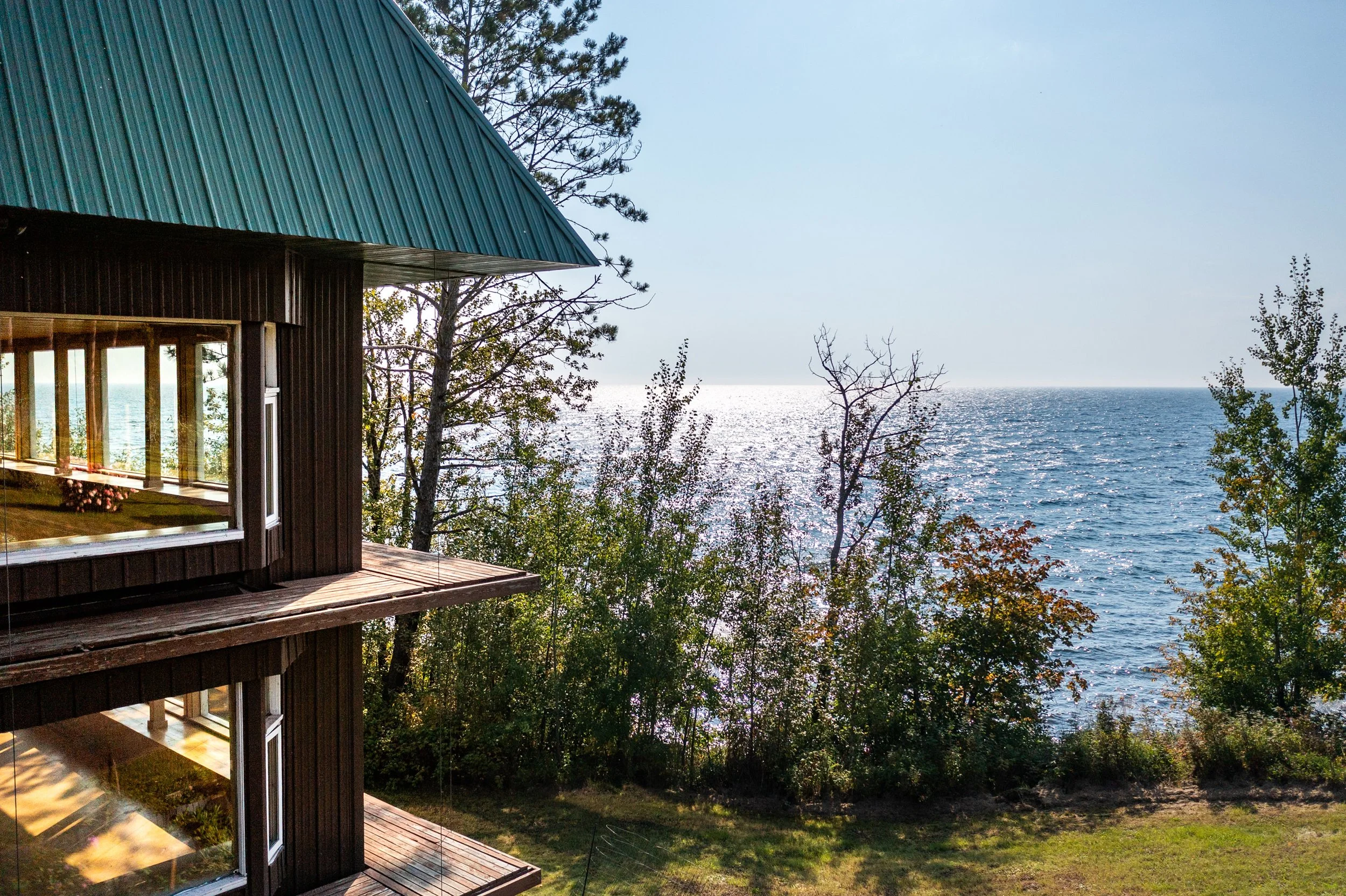 A wooden house with a teal metal roof overlooking a body of water, with trees and foliage in the foreground.