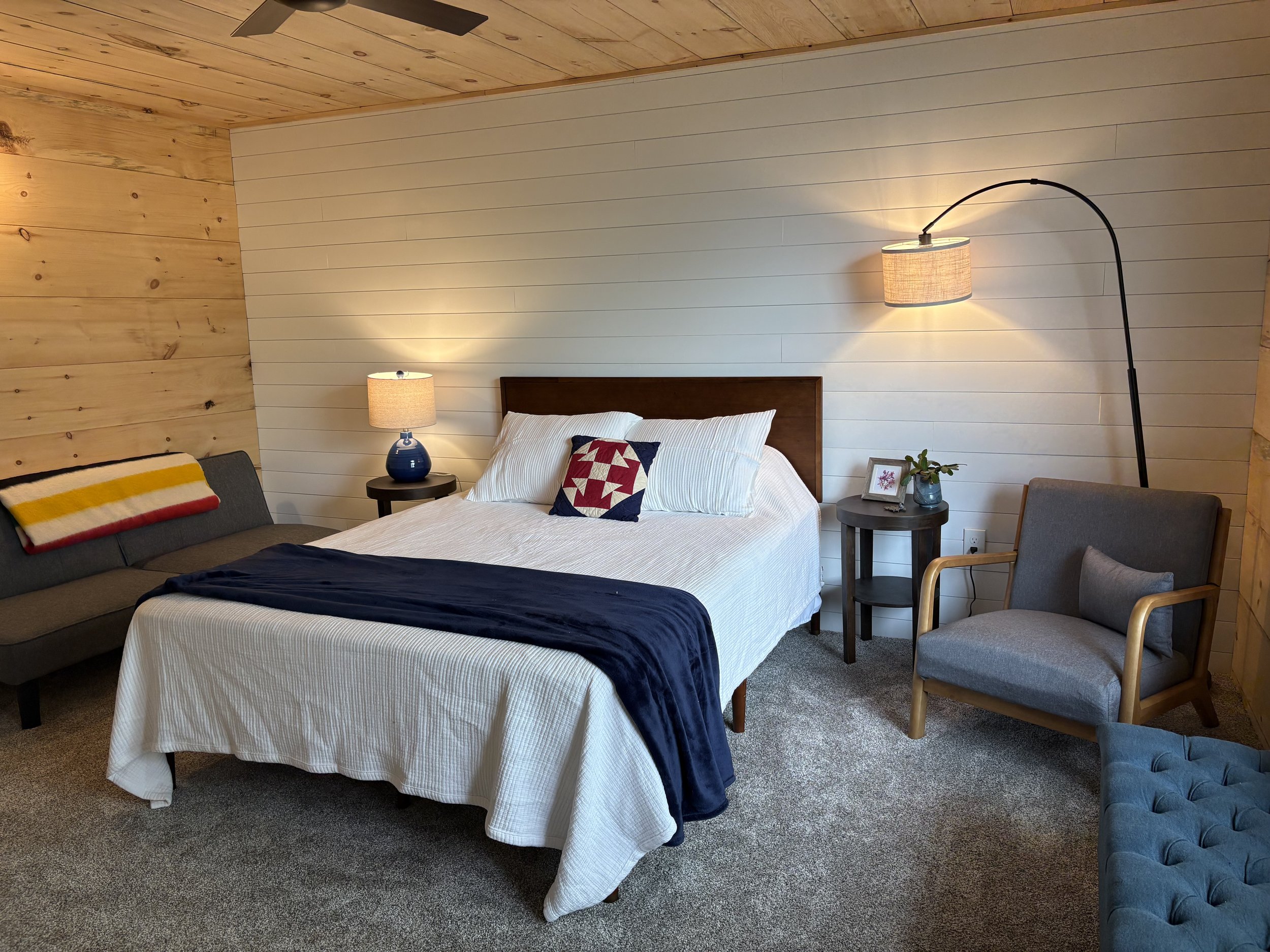 A bedroom with a wooden-paneled ceiling and walls. It has a bed with white bedding and a geometric pillow, a bedside table with a blue lamp, a sitting area with a gray sofa and an armchair, a black floor lamp with a beige lampshade, and a small plant