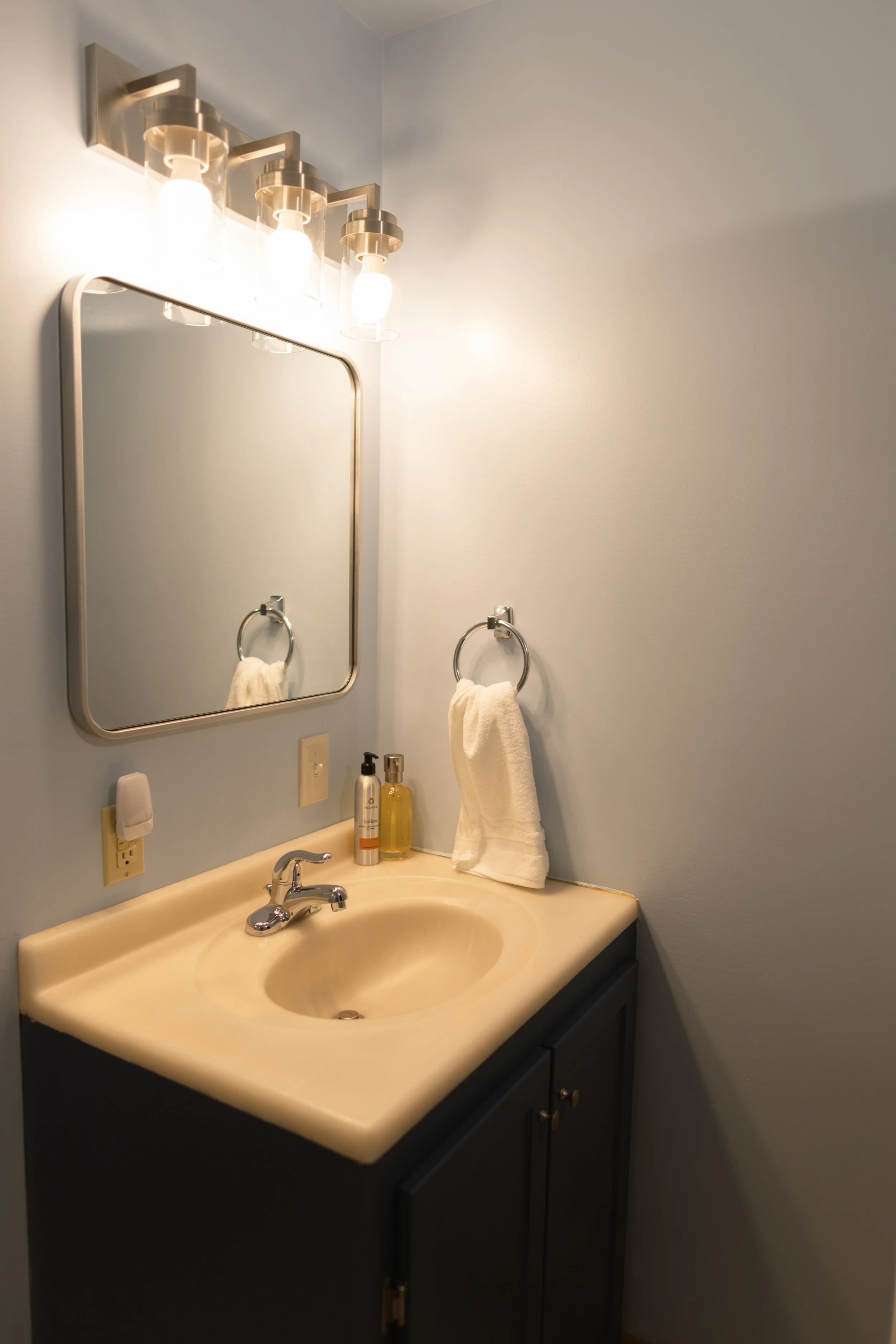 Bathroom vanity with a beige sink, a silver faucet, a dark cabinet with gold knobs, a mirror, a wall-mounted towel ring with a white towel, wall-mounted soap and lotion bottles, and a light fixture with three exposed bulb lights.