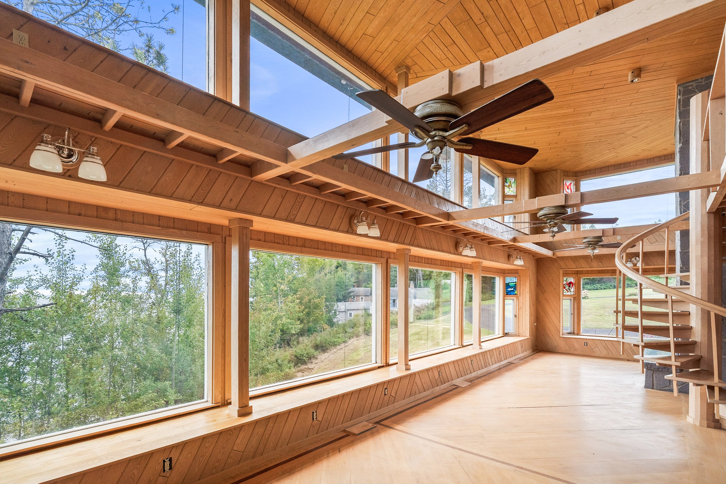 Interior of a wooden house with large windows, ceiling fans, and a spiral staircase, overlooking greenery and a clear blue sky outside.