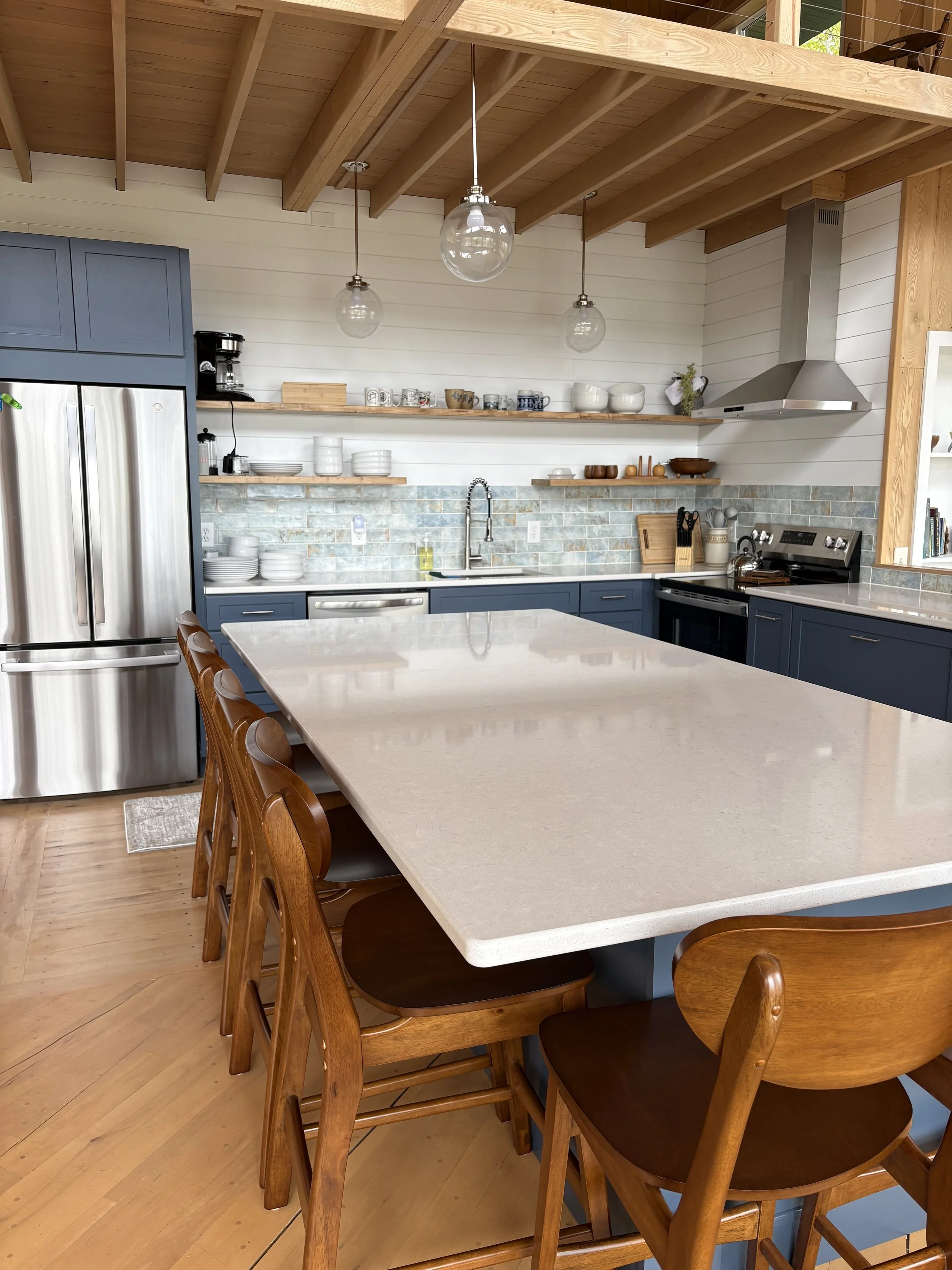 A modern kitchen with a large off-white island, wooden chairs, blue cabinets, stainless steel refrigerator, open wooden shelves with dishes and cookware, a black stove, and pendant lights hanging from a wooden ceiling.