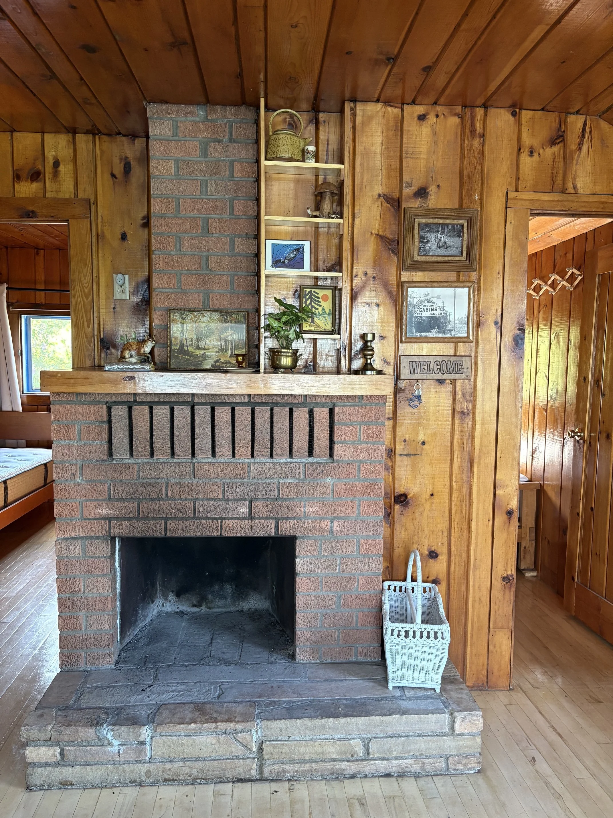 A fireplace with a brick and stone base, a wooden mantel, and a wooden wall behind it. Decorative items on the mantel include a horse sculpture and framed pictures. To the right, there is a white wicker basket on the floor, and a doorway leading to a