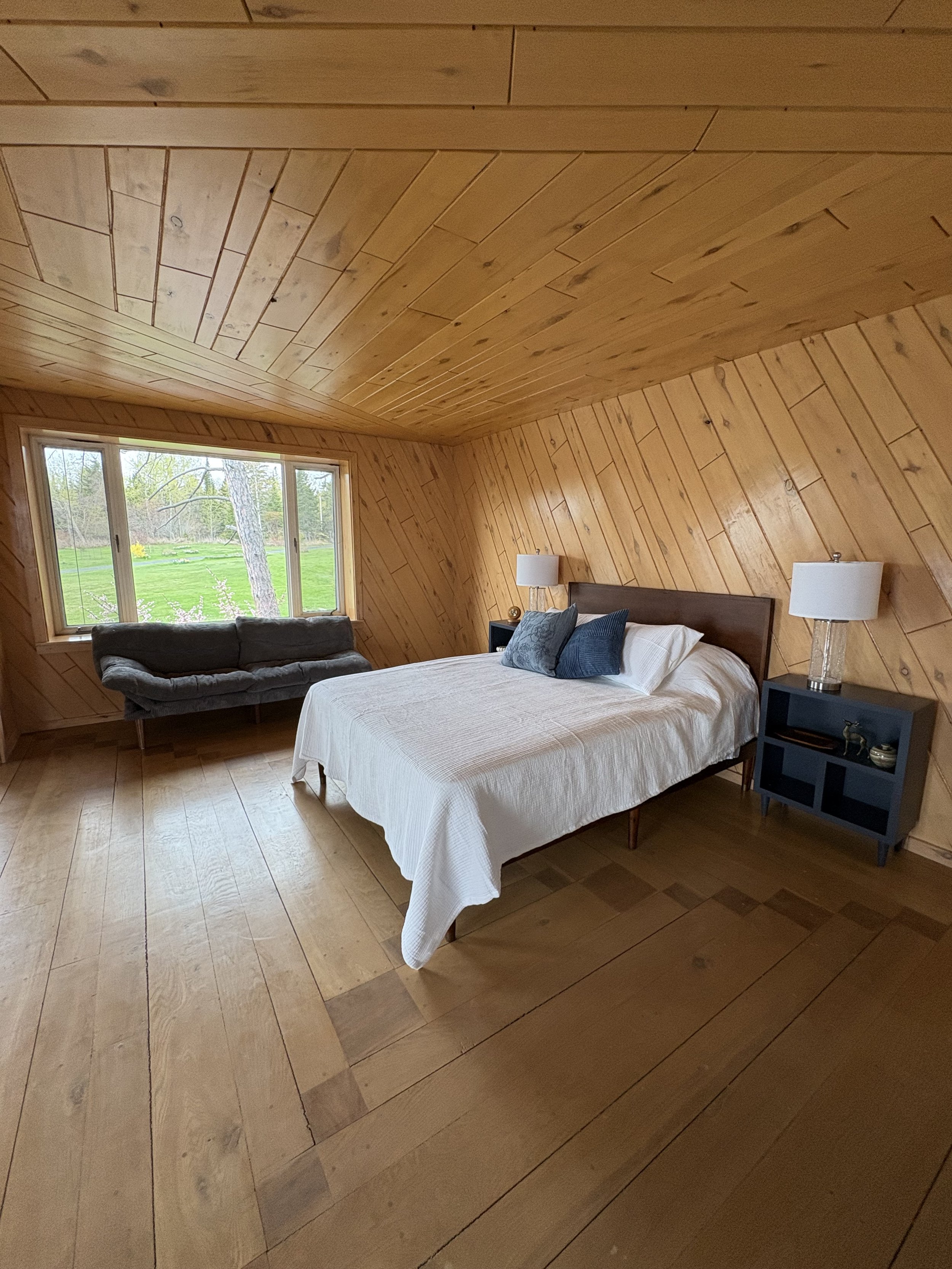 A bedroom with wooden paneled walls and ceiling, a window showing green outdoor scenery, a bed with white bedding and blue pillows, two bedside tables with white lamps, and a dark gray armchair at the foot of the bed.