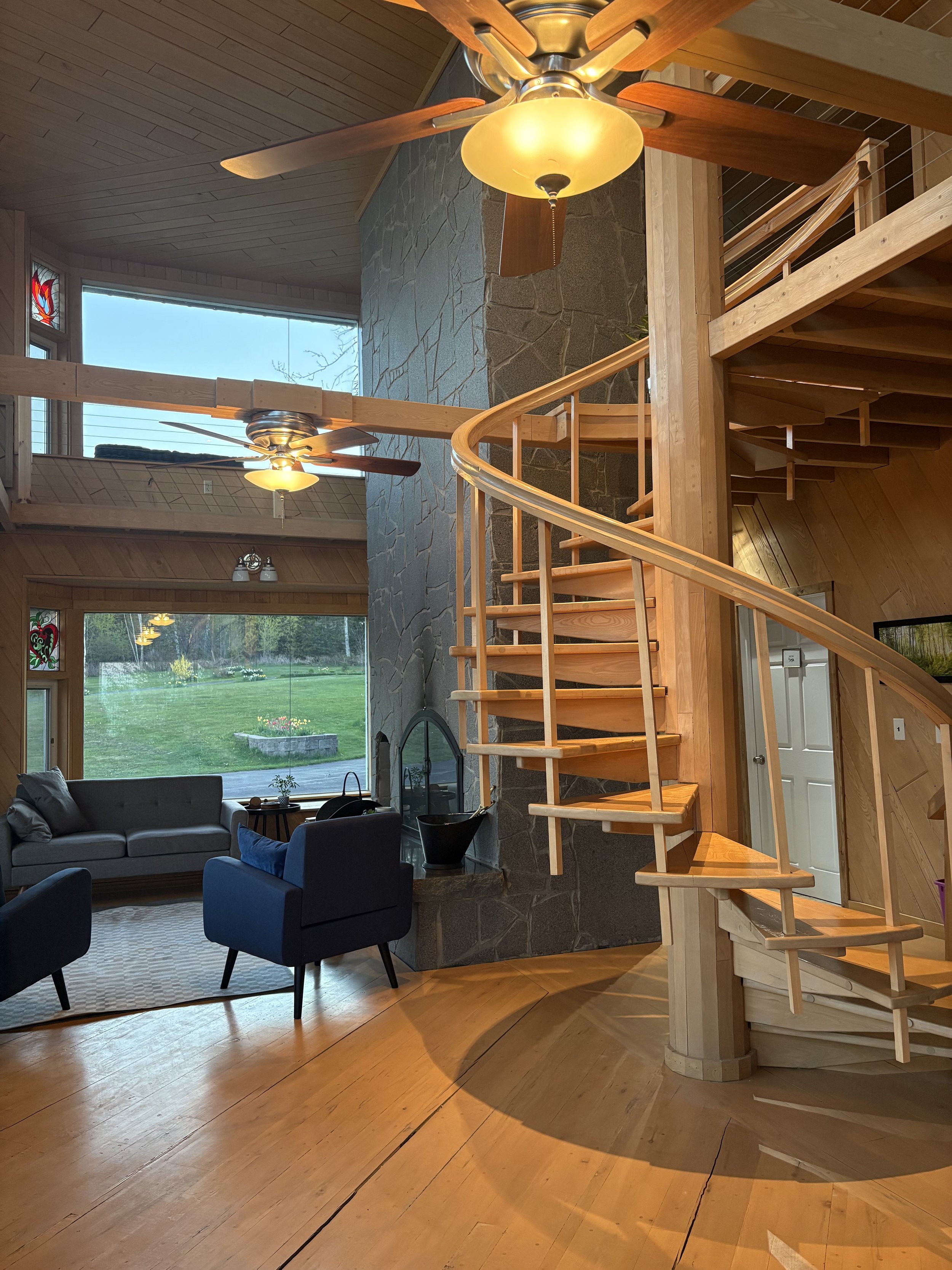 Interior view of a house with a wooden spiral staircase, a stone fireplace, large windows showing a green lawn, and wooden flooring.