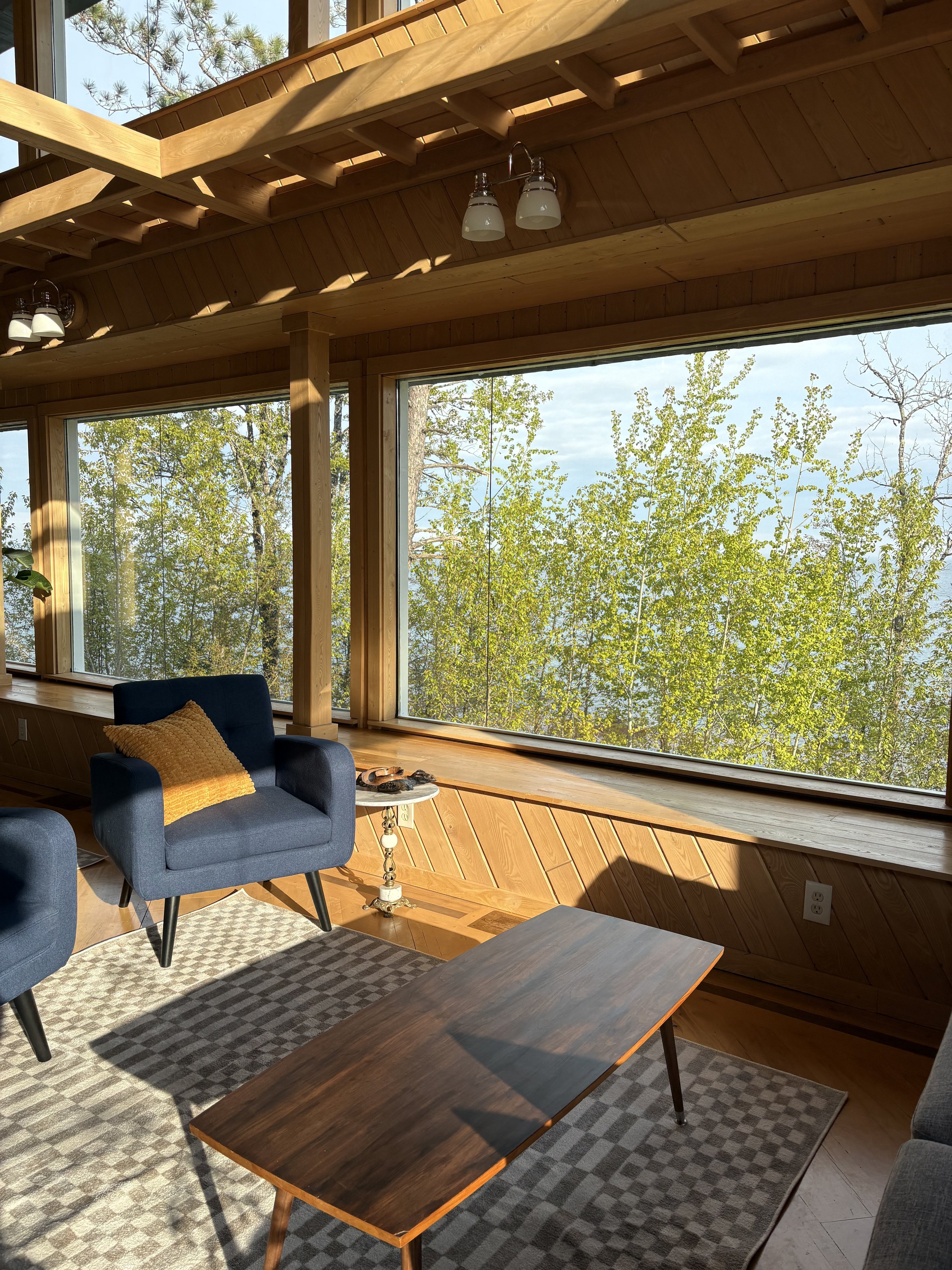 Interior of a house with large windows showing green trees outside. There is a navy blue armchair with a yellow cushion, a patterned area rug, and wooden tables and wall paneling, with sunlight streaming in.
