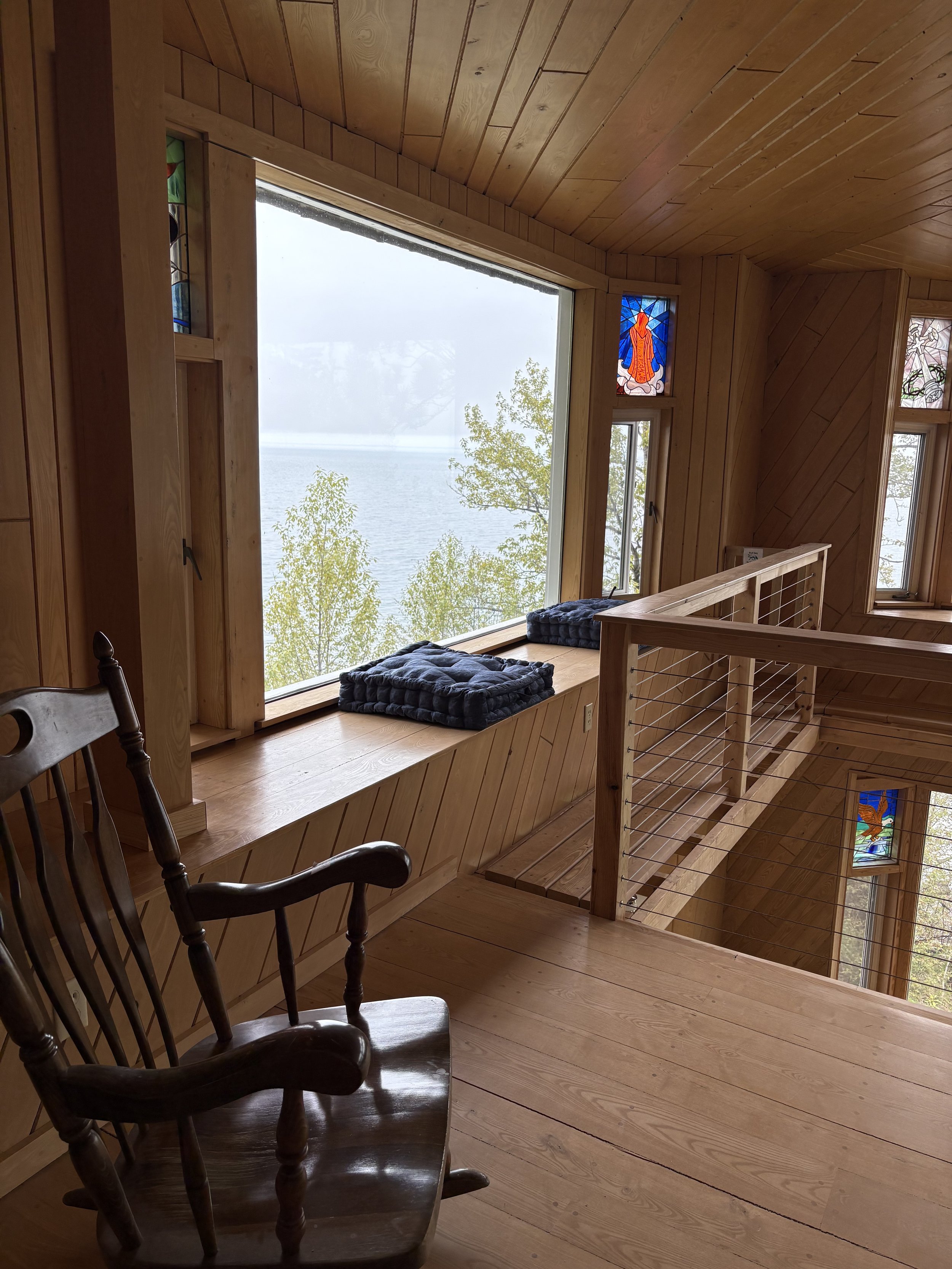 Interior of a wooden house with large windows showing a foggy lake and trees outside. Two stained glass windows depict spiritual figures, and a wooden bench features folded blankets. A wooden chair is partially visible in the foreground.