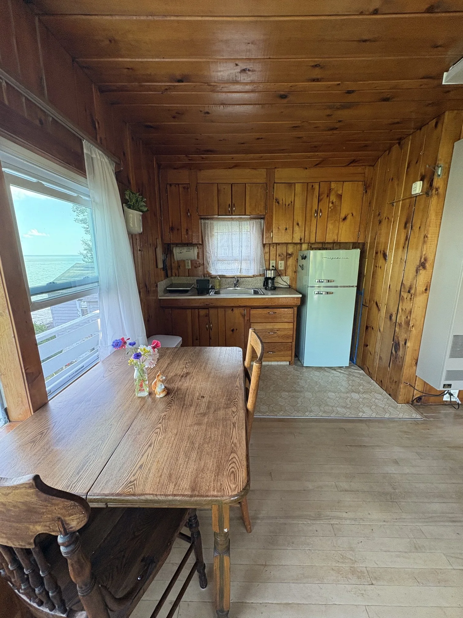 A rustic kitchen with wood-paneled walls and ceiling, a window with white curtains, a table with a vase of colorful flowers, and a partial view of wooden chairs.