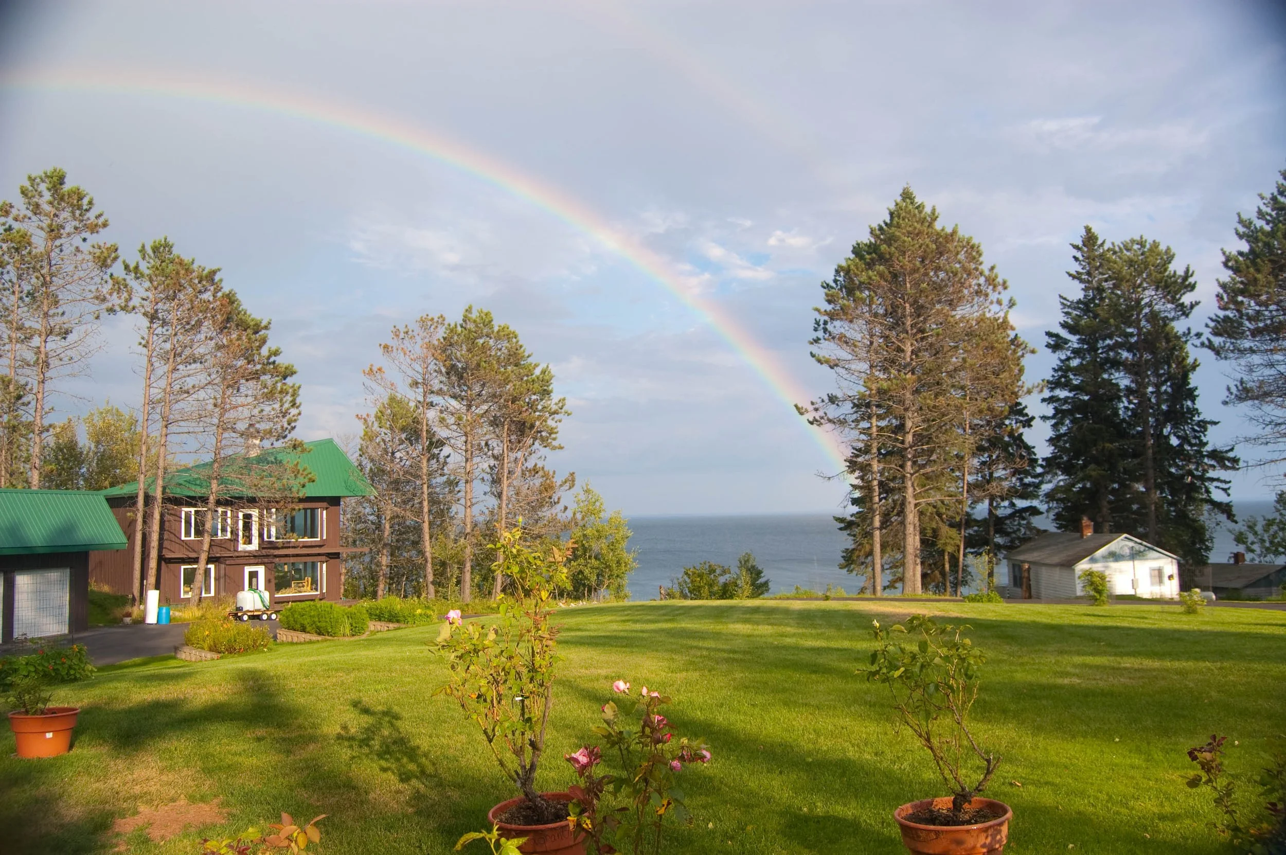 View of a rainbow arching over a residential area with trees, houses, and a body of water in the background, on a partly cloudy day.