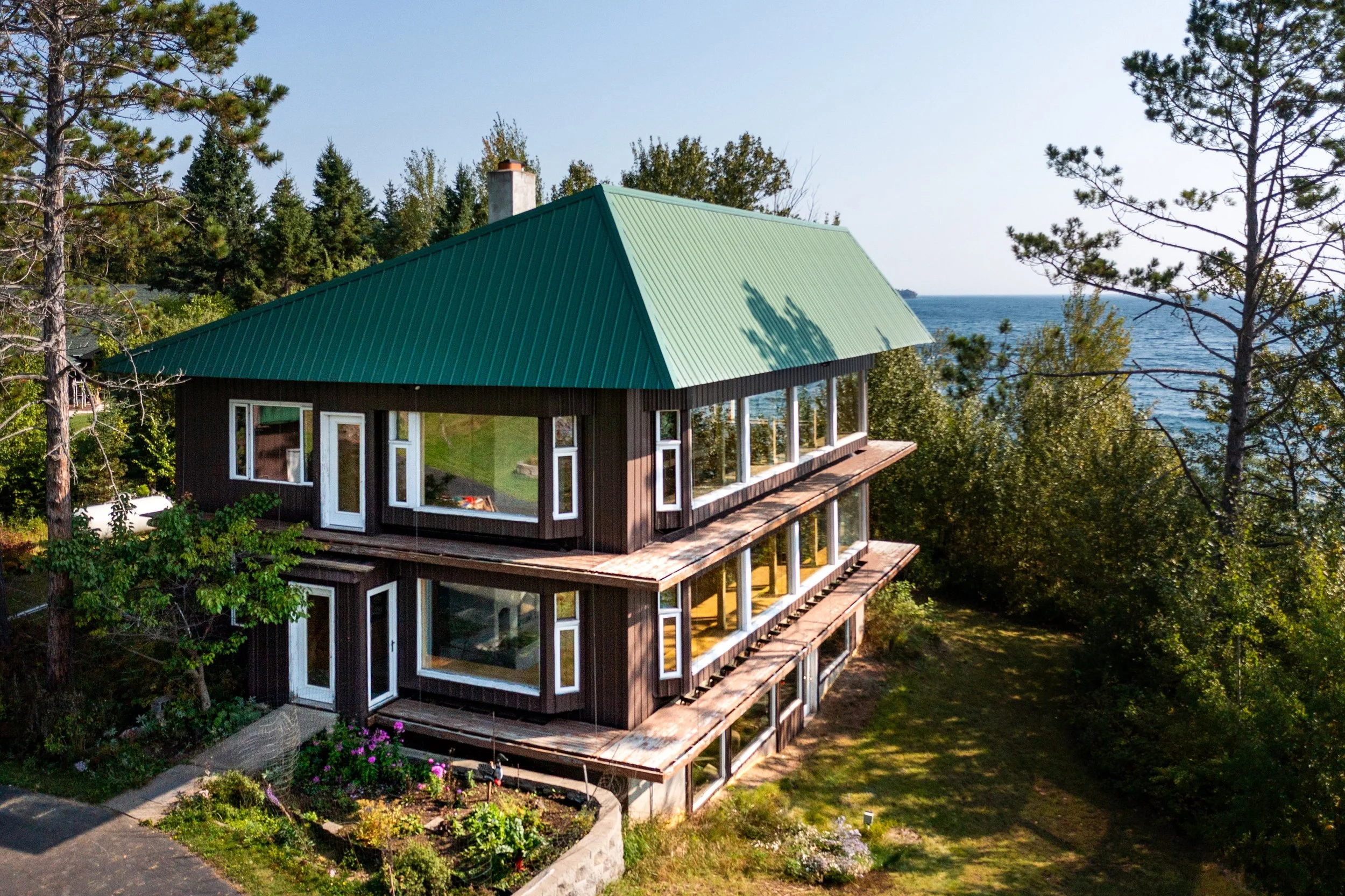 A multi-story house with a green metal roof and large windows, situated among trees overlooking a body of water under a clear sky.