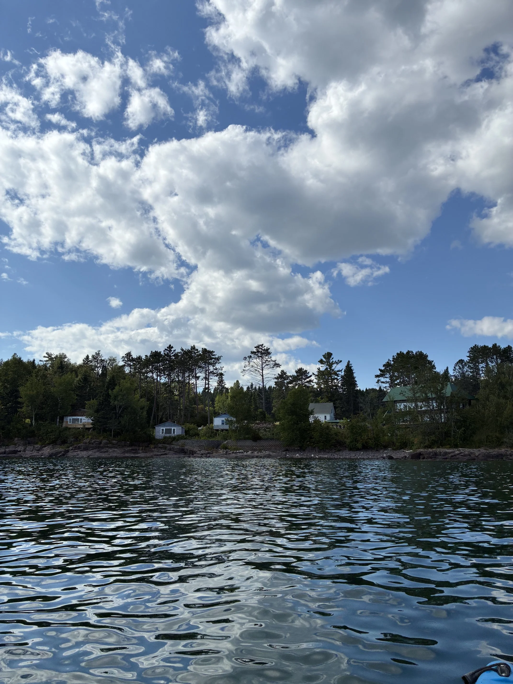View of a body of water with gentle ripples, a shoreline with trees, and houses with green roofs in the background under a partly cloudy sky.